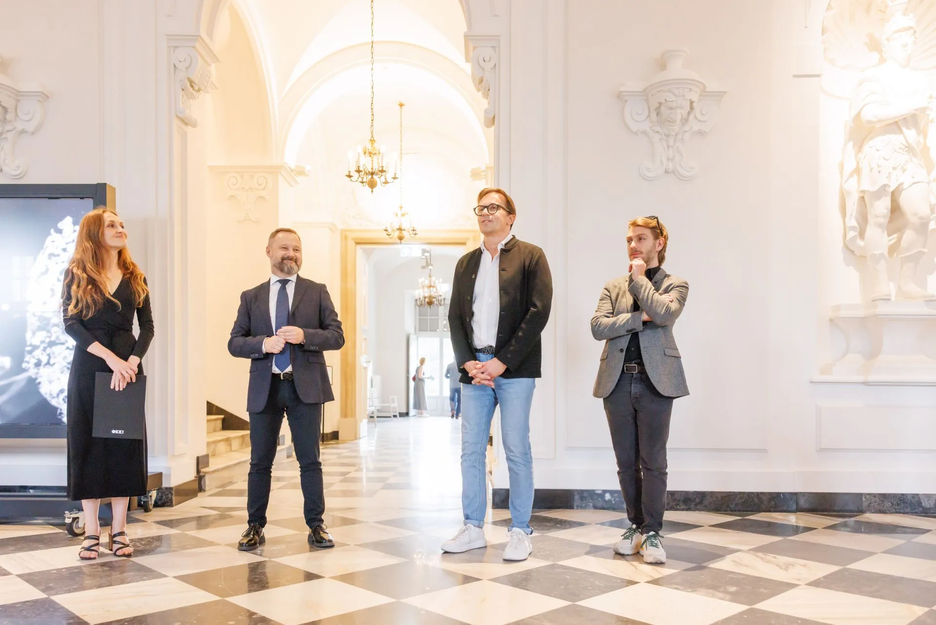 Four people standing in a grand hall with white walls, chandeliers, and a checkered floor