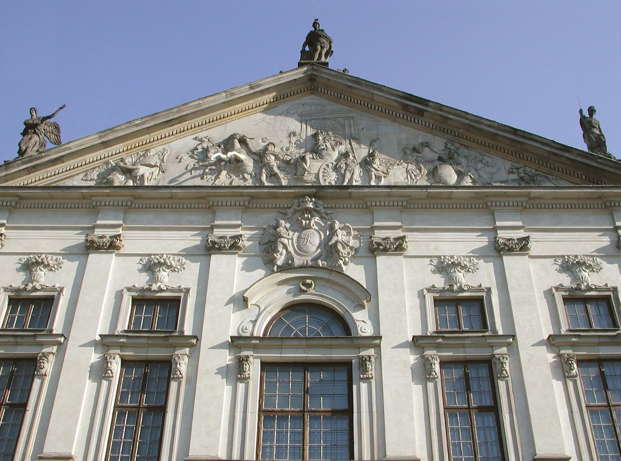 Barokke paleisgevel met wit stucwerk, rondboogvensters en een gedetailleerd sculpturaal fronton onder een helderblauwe hemel