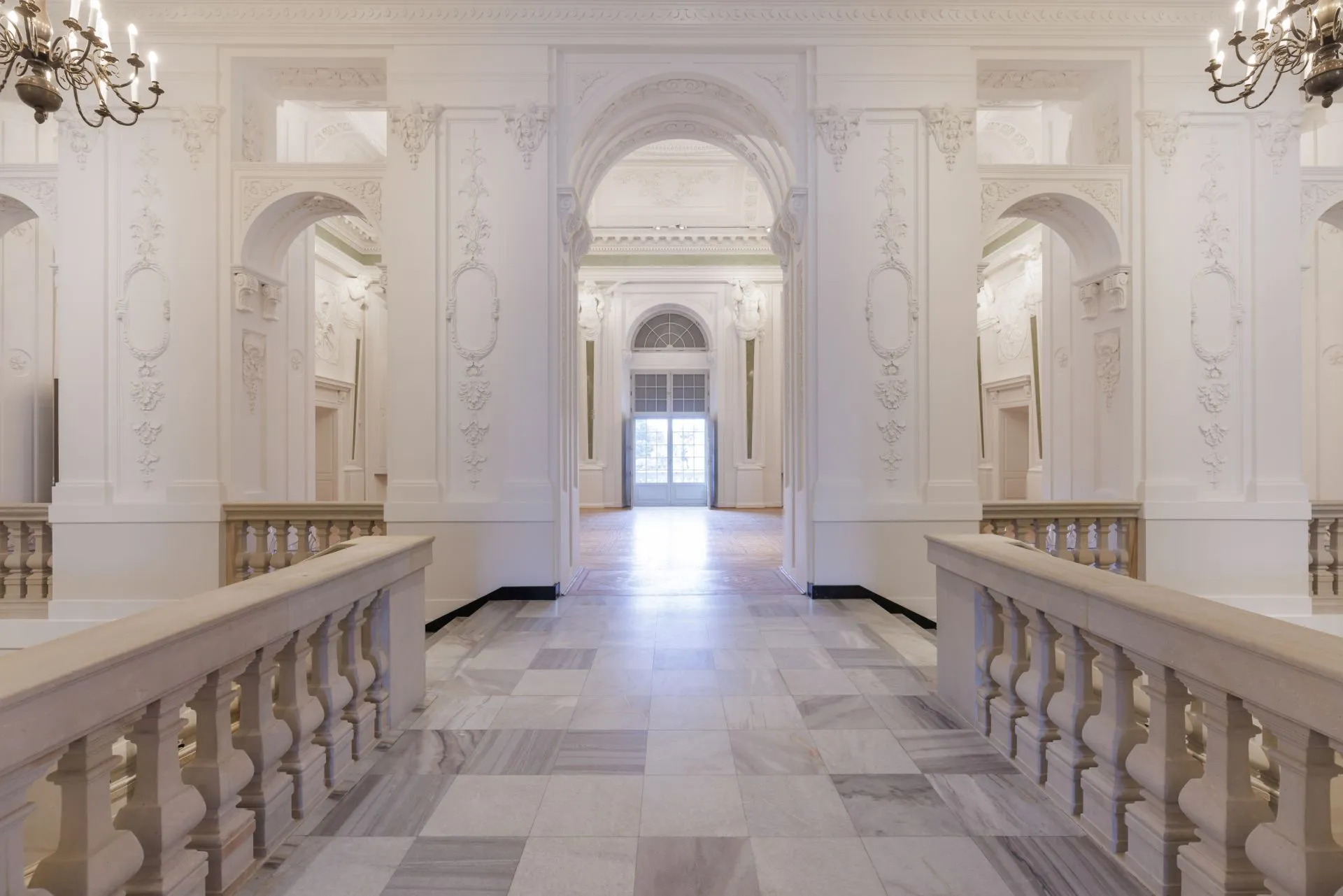 Een lange gang met een witte marmeren vloer, sierlijke balustrades en boogvormige deuropeningen in het interieur van een historisch paleis.