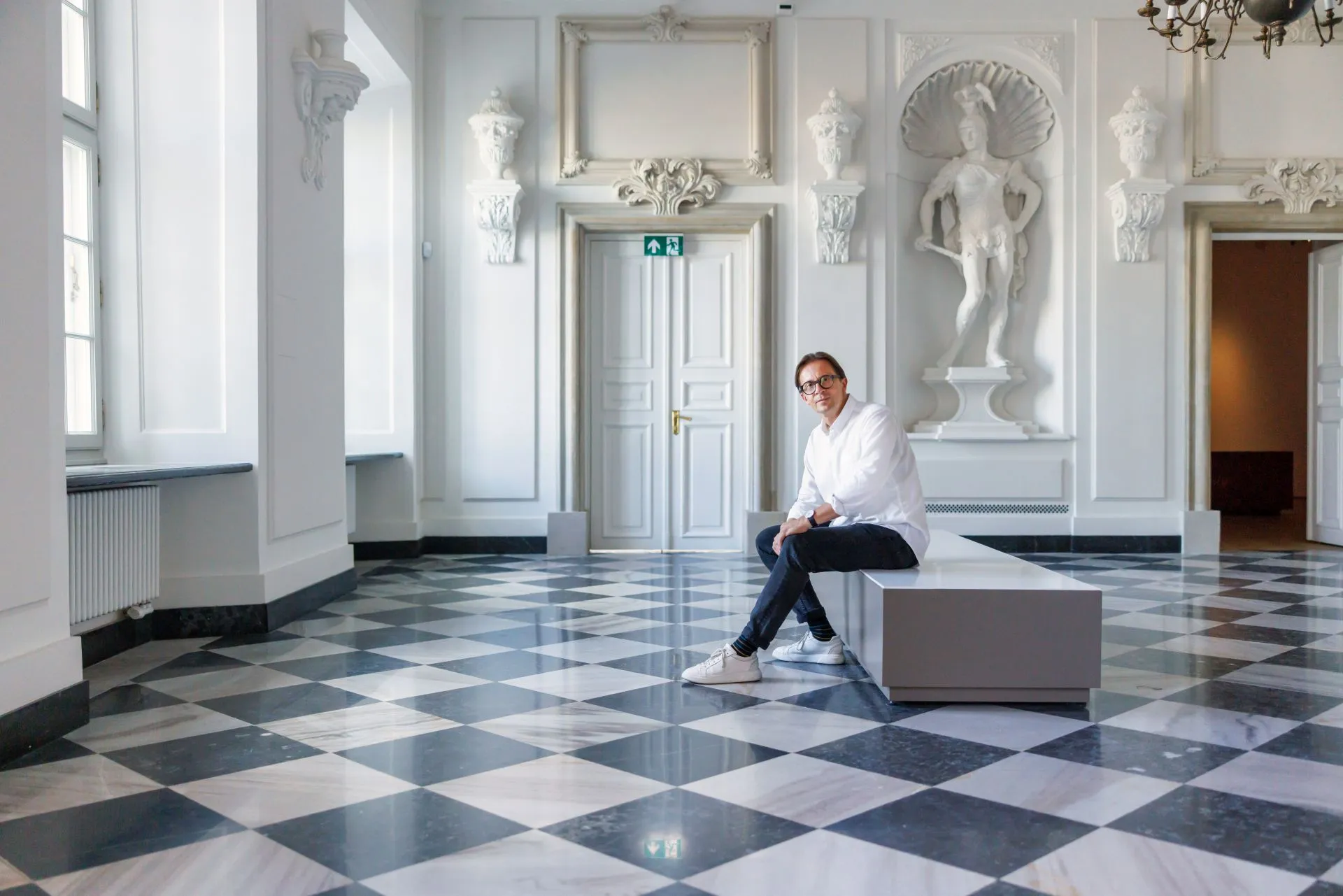 A person sitting on a bench in a grand hall with white walls, a black and white checkered floor, and classical sculptures.