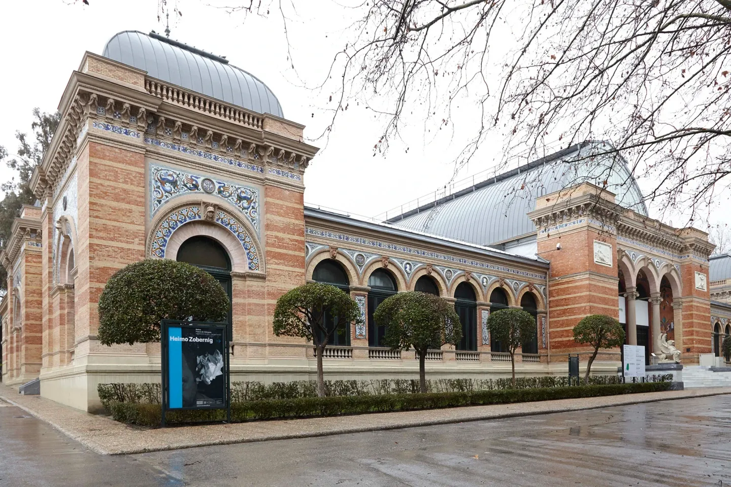 Zewnętrzna część Palacio de Velázquez z ceglaną fasadą, łukowymi oknami i kopułowym dachem, otoczona przyciętymi żywopłotami i nagimi drzewami.