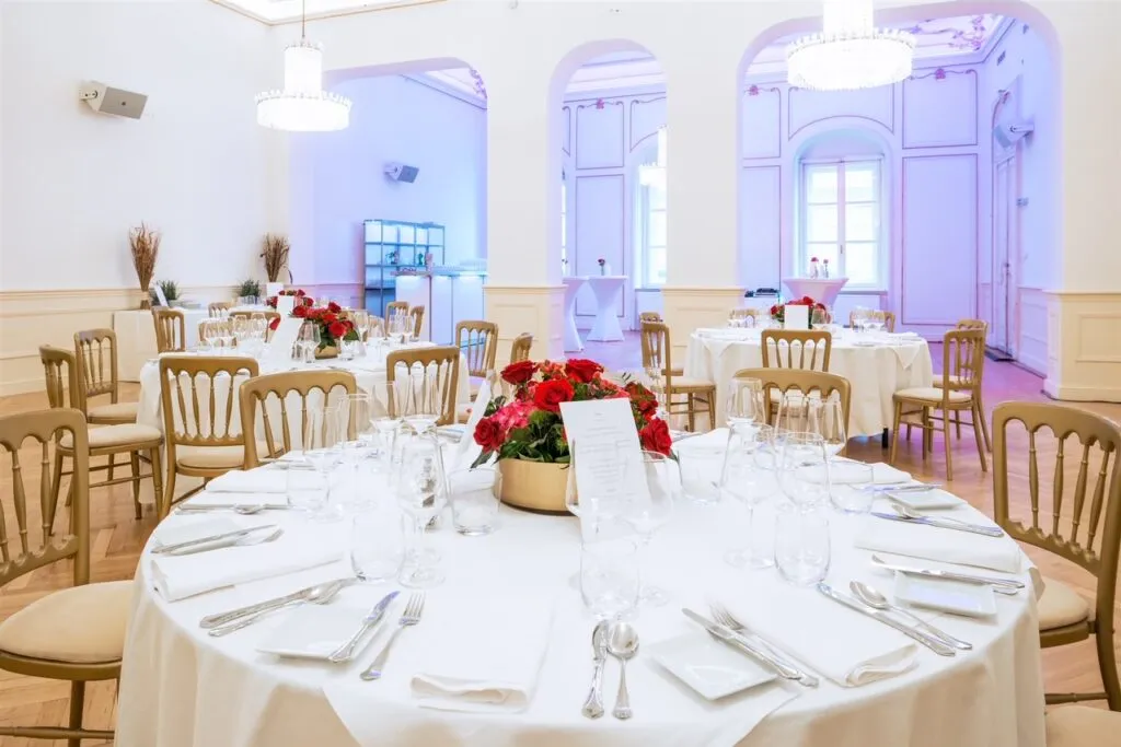 Ronde tafel gedekt met wit linnen, bestek en rode bloemen in een statige kamer met boogramen en kroonluchters