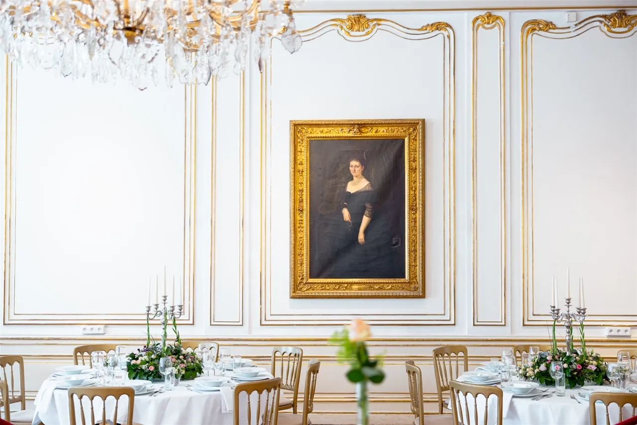 Barok interieur met wit geprofileerde wanden, gouden randen, een kroonluchter en een ingelijst portret boven een eettafel.