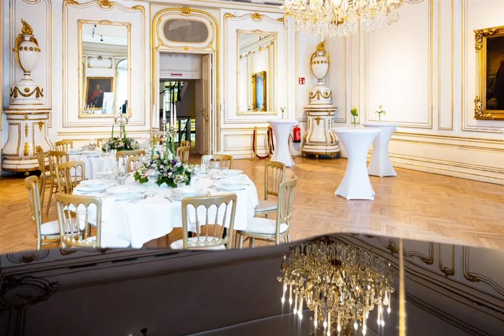 Ornate barok interieur met witte muren, gouden randen, spiegels en een kristallen kroonluchter, met een ronde tafel gedekt voor een evenement.
