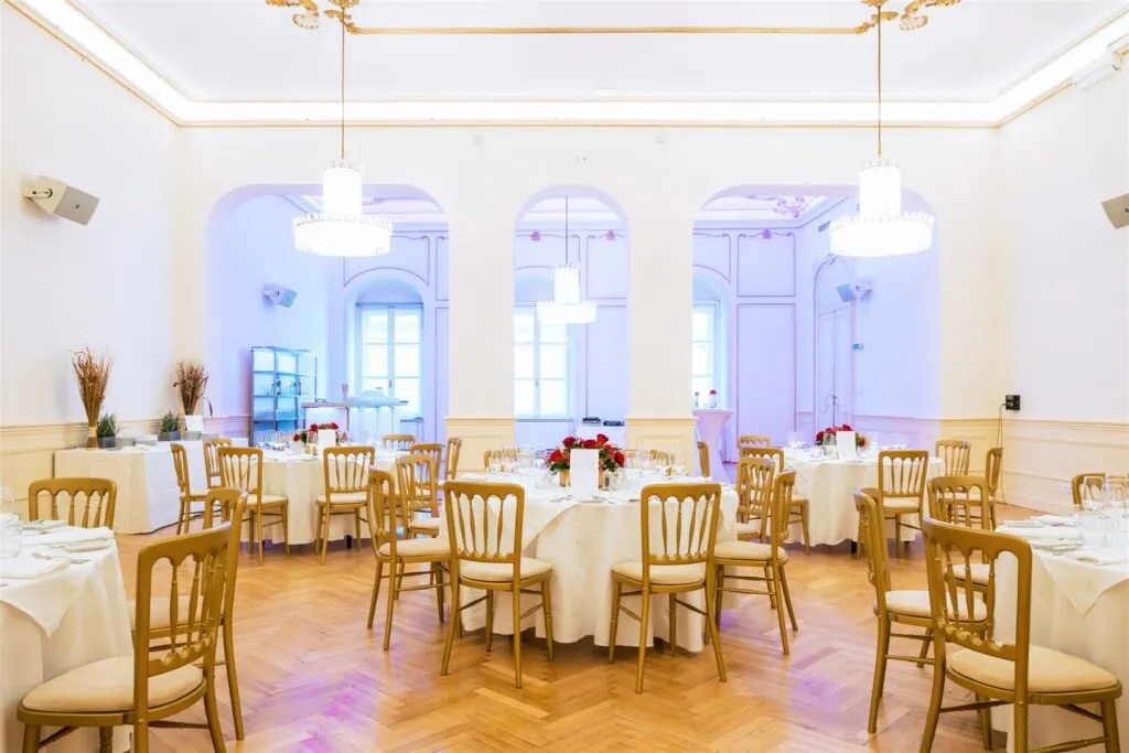 Een ruime balzaal met witte tafels, houten stoelen en kroonluchters onder hoge plafonds.