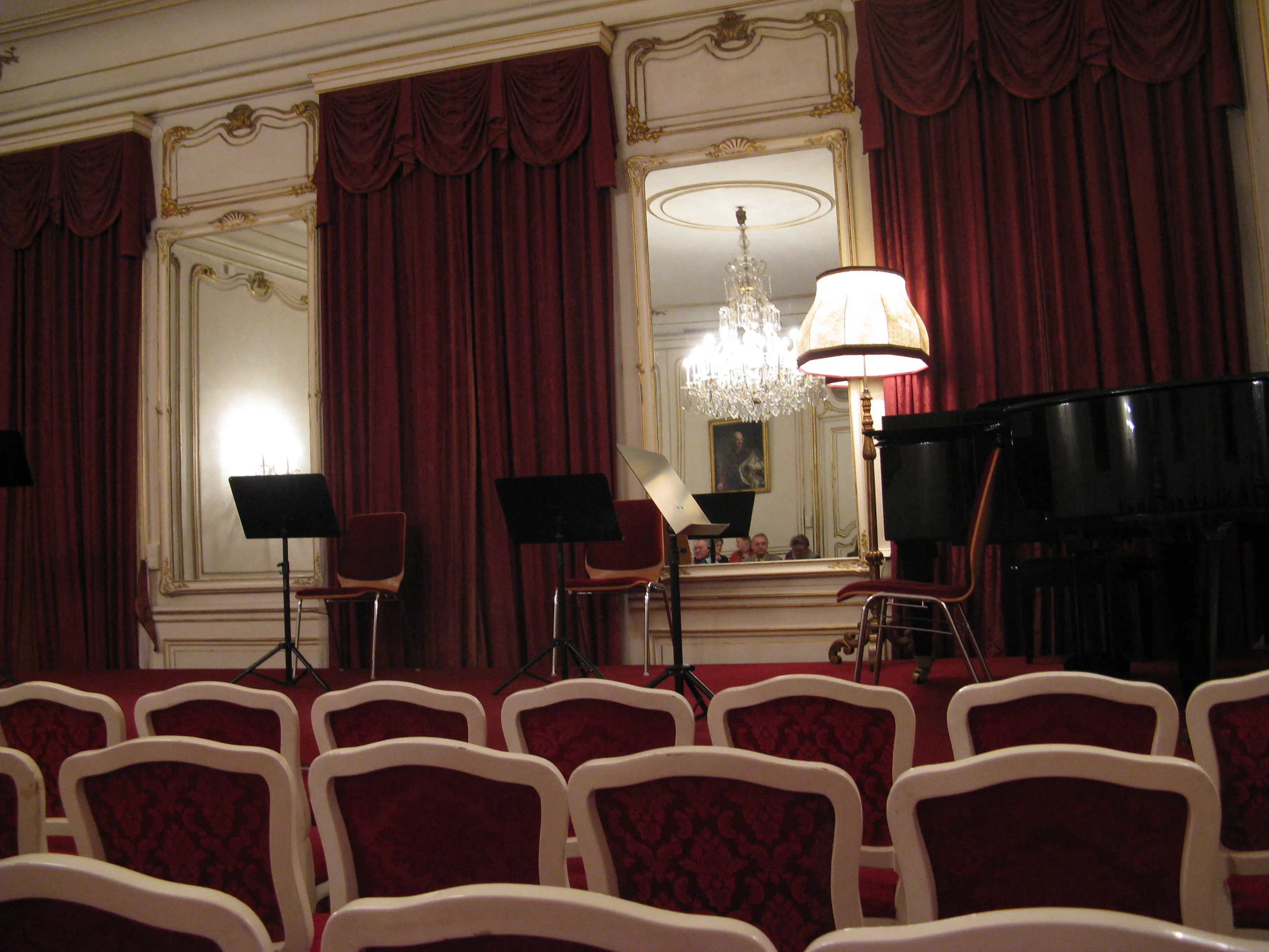 Barok interieur met rood fluwelen gordijnen, witte sierlijke ornamenten, een vleugel en rijen rood gestoffeerde stoelen in een concertzaal