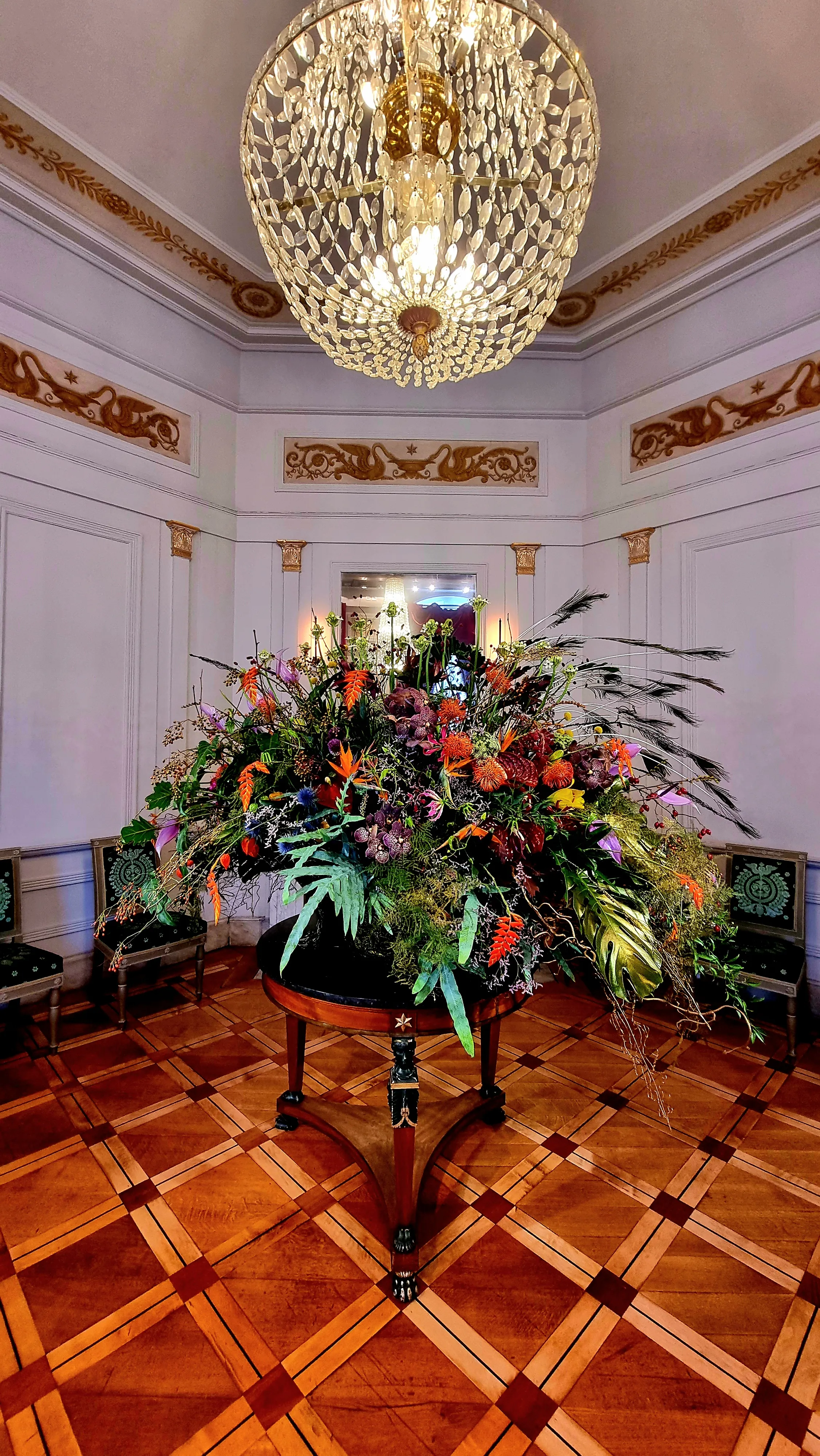 Gran arreglo floral colorido sobre una mesa redonda bajo una lámpara de araña de cristal en una ornamentada sala de palacio con suelos de parqué y frisos dorados en las paredes