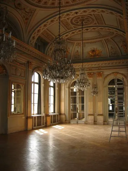 Interior de un gran salón de baile con techo ornamentado y pintado, lámparas de cristal, ventanas arqueadas y suelo de madera pulida