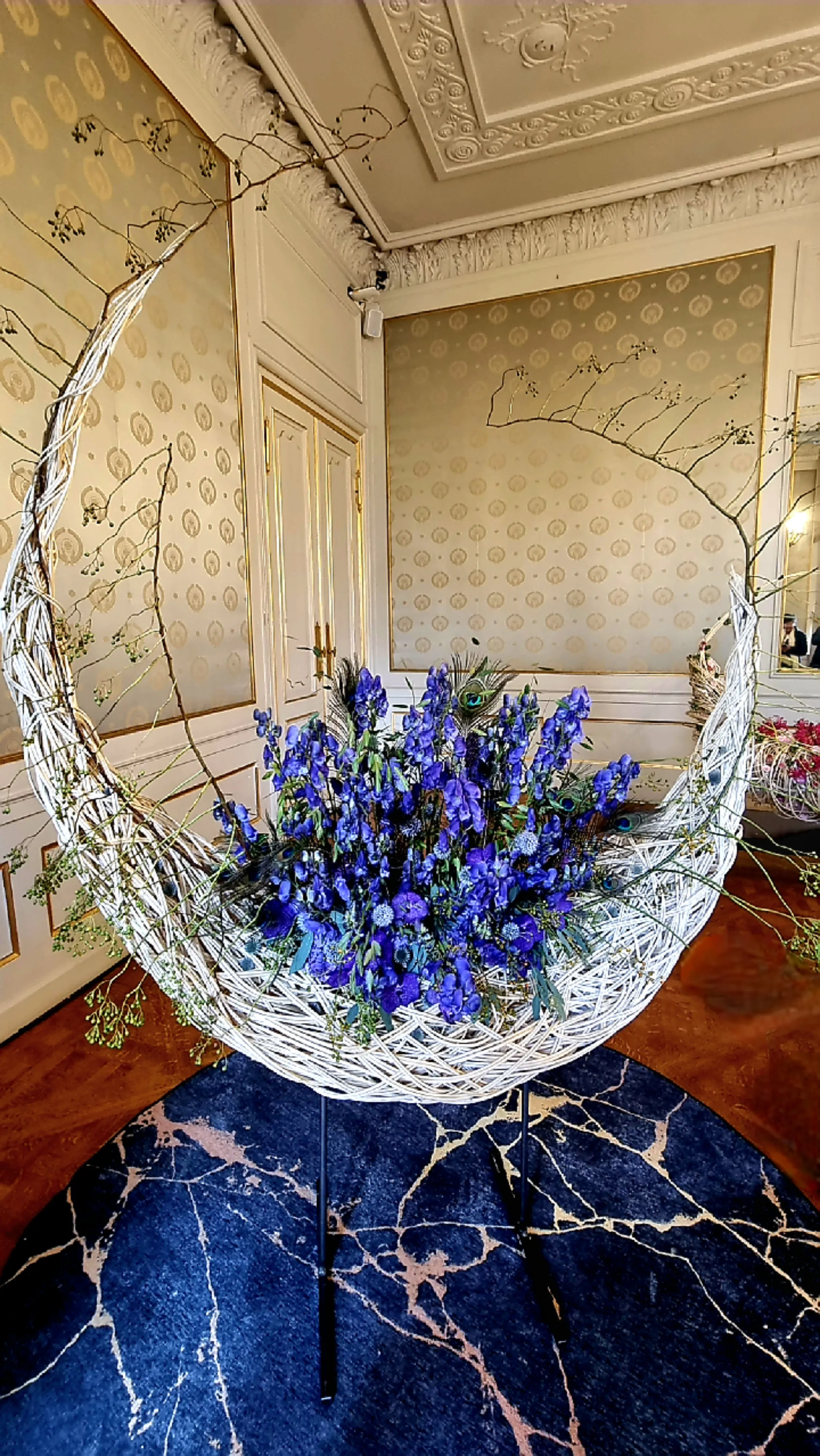 Een groot, halvemaanvormig wit rieten beeldhouwwerk gevuld met blauwe bloemen en pauwenveren staat op een blauw met goudkleurig tapijt in een rijk versierde Rococo-kamer met behang met patronen en decoratief lijstwerk aan het plafond.