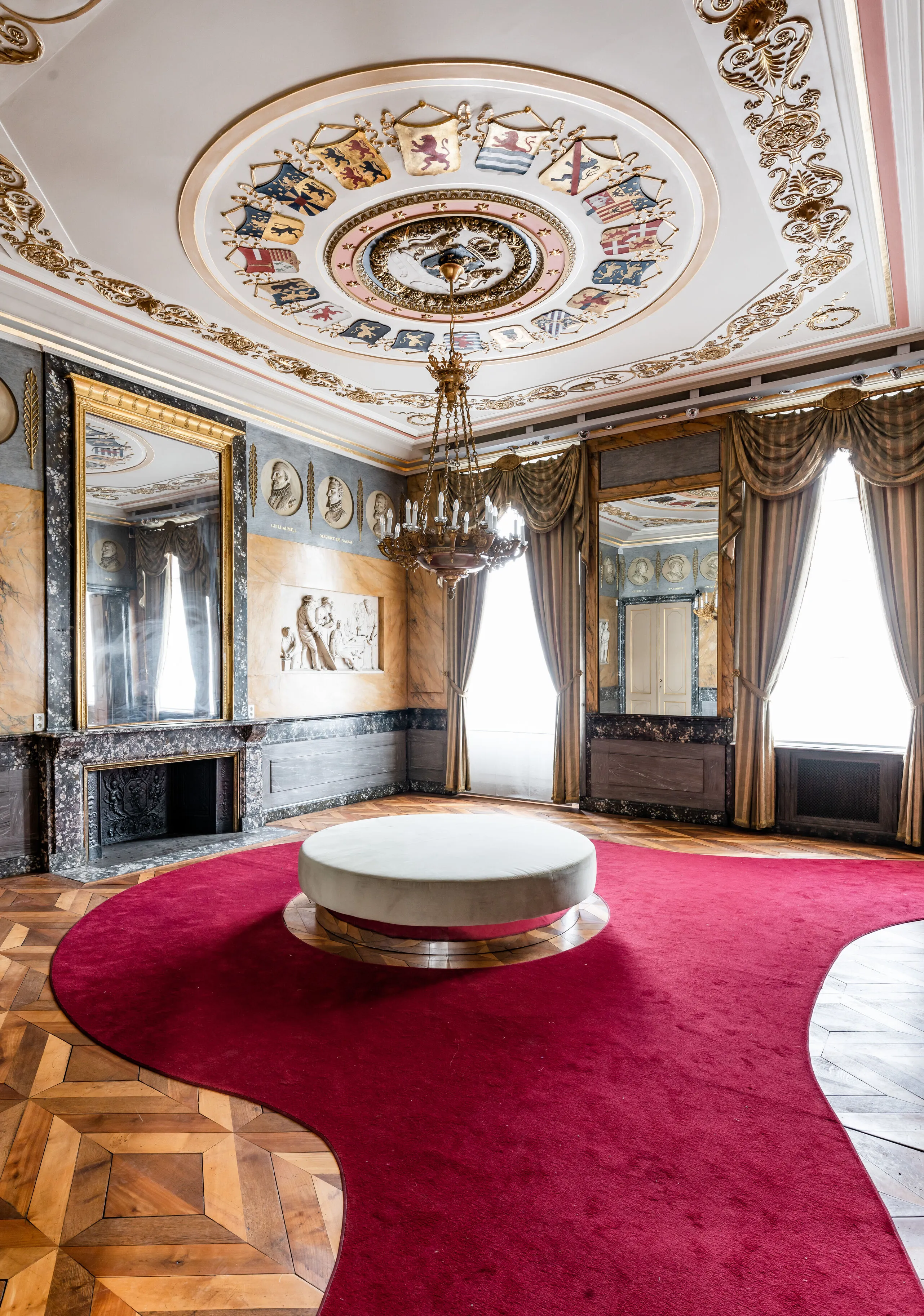 Ornado salón rococó con un techo pintado con escudos de armas, una lámpara de araña, grandes espejos, chimenea, suelo de parqué y una alfombra roja con un otomán redondo.