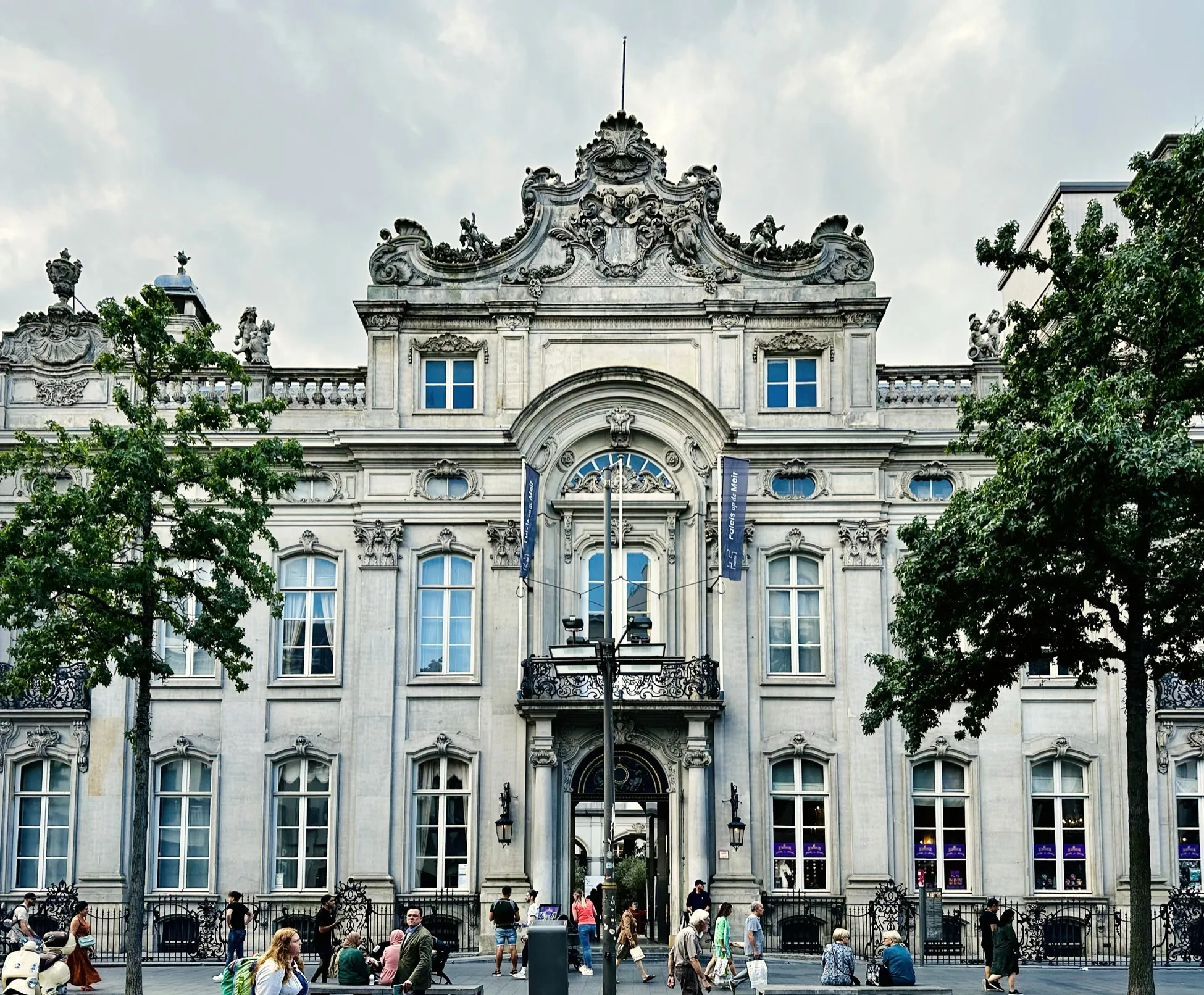 Fachada rococó ornamentada del palacio Paleis op de Meir en Amberes con una entrada central arqueada, cantería decorativa, banderas azules y visitantes reunidos en la plaza de abajo
