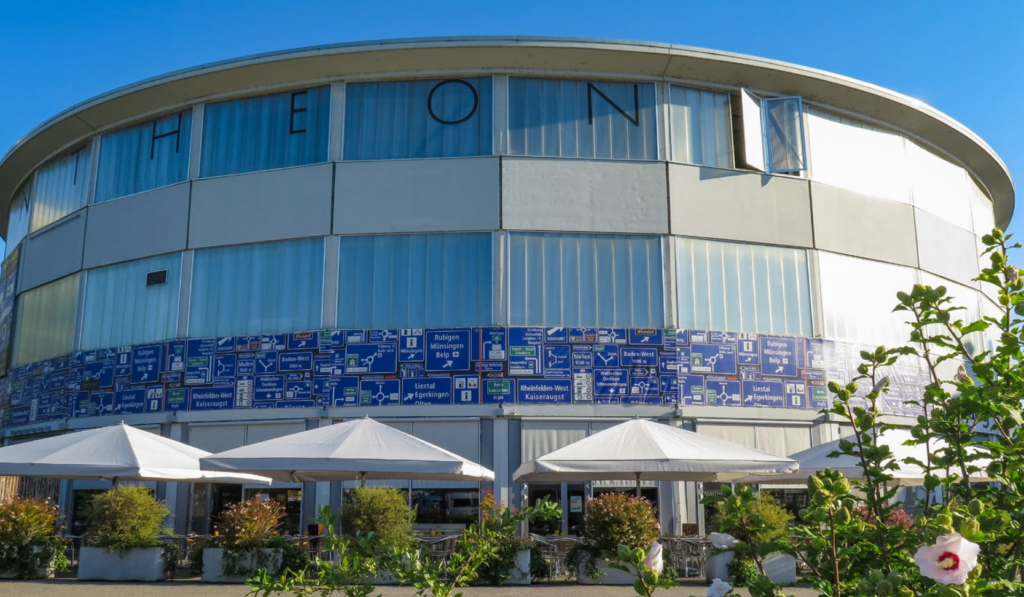 Fachada circular de un edificio con la inscripción PANTHEON sobre las ventanas superiores, sombrillas blancas y vegetación en primer plano