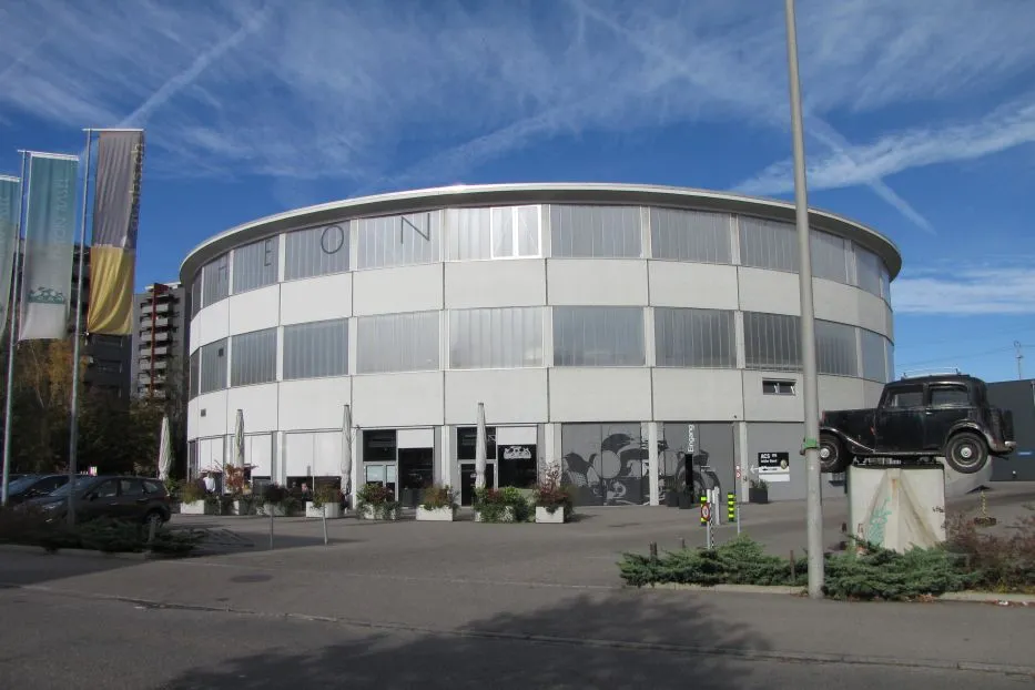 Edificio de museo redondo de color blanco y gris con bandas de ventanas horizontales y un coche negro antiguo expuesto sobre un pedestal en el exterior