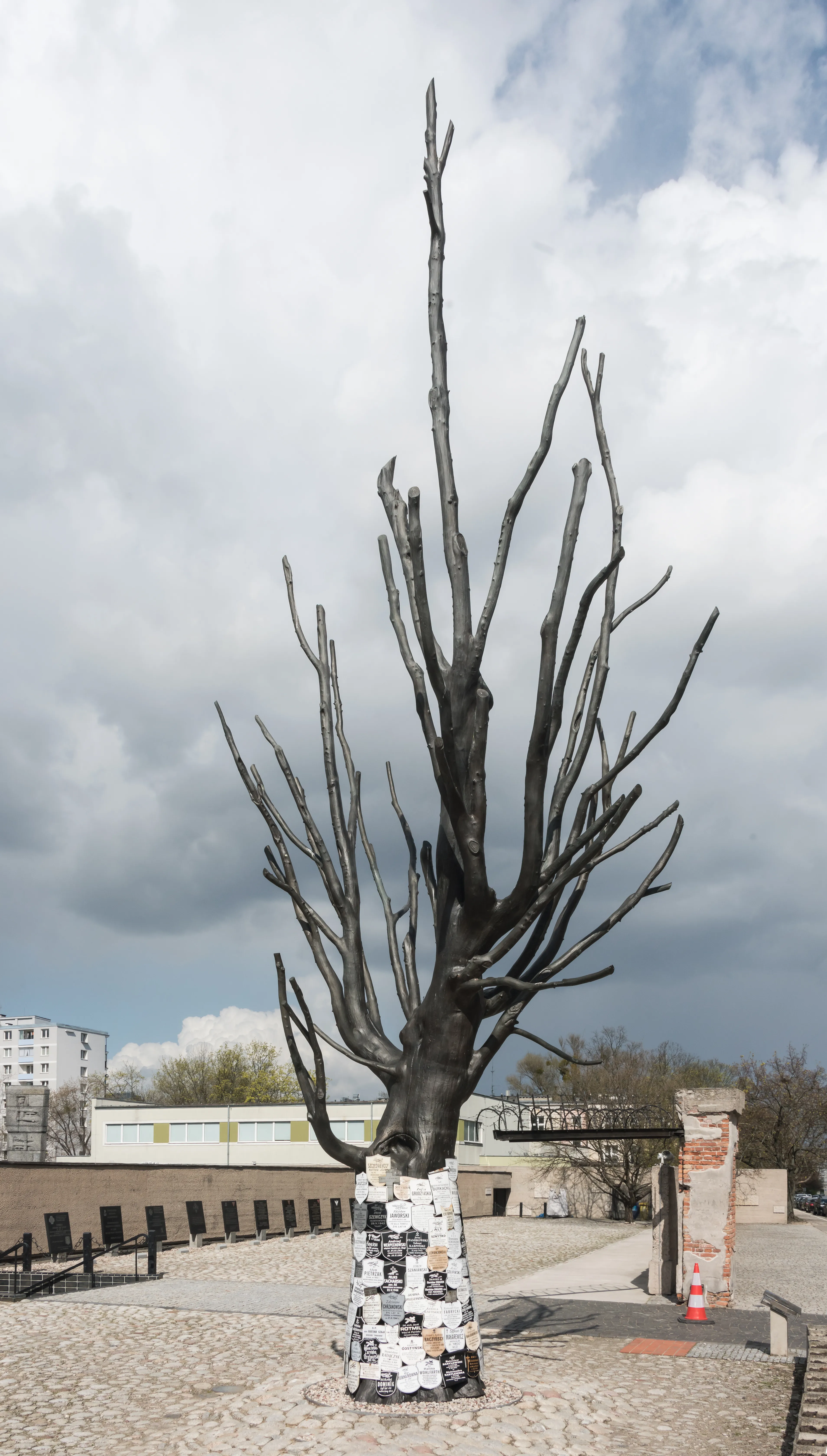 A large dark metal sculpture of a bare tree with a base covered in white tiles with black inscriptions, set in an outdoor courtyard with buildings and a cloudy sky in the background.