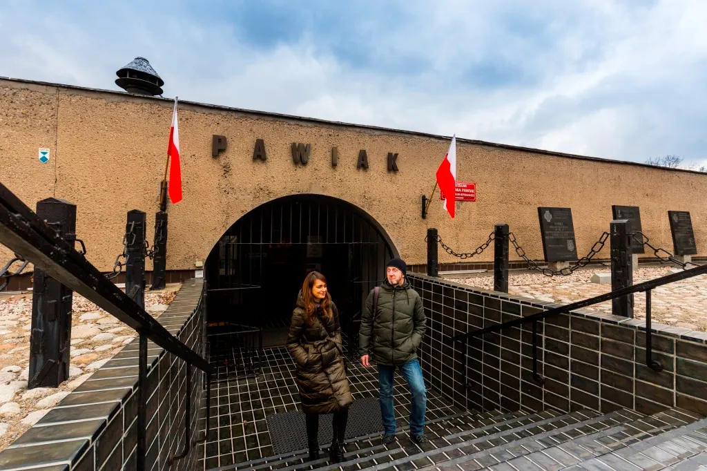 Two people standing at the entrance of Pawiak Prison Museum, with a large arched gateway and the name 'PAWIAK' on the building facade.