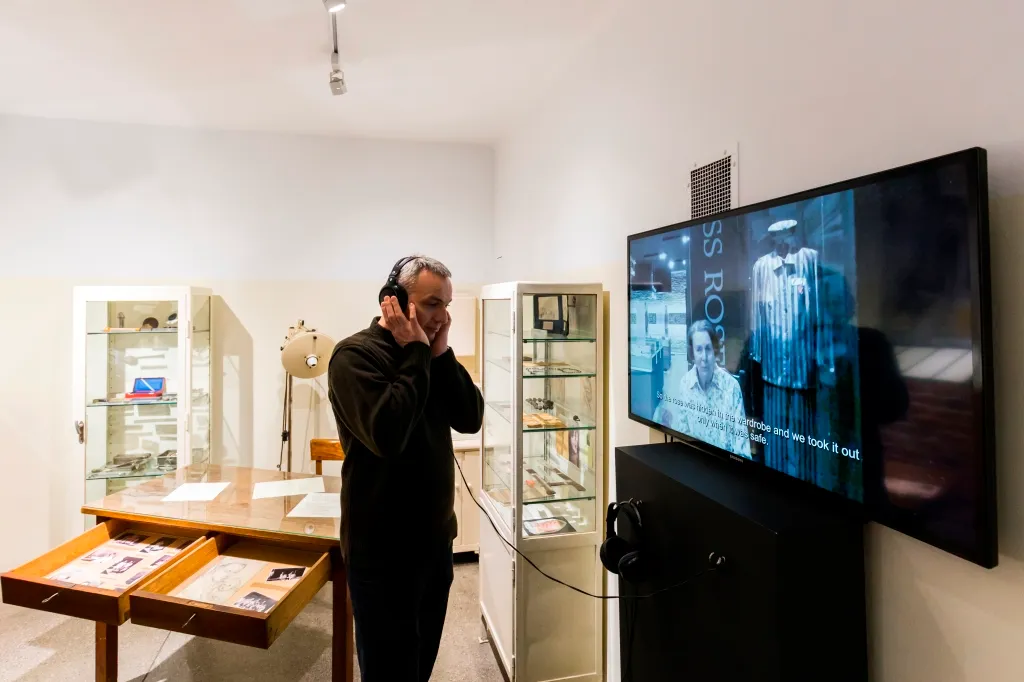 A man wearing headphones stands in a museum gallery, looking at a large screen displaying a historical photograph and text, with glass display cases and a wooden table in the background.
