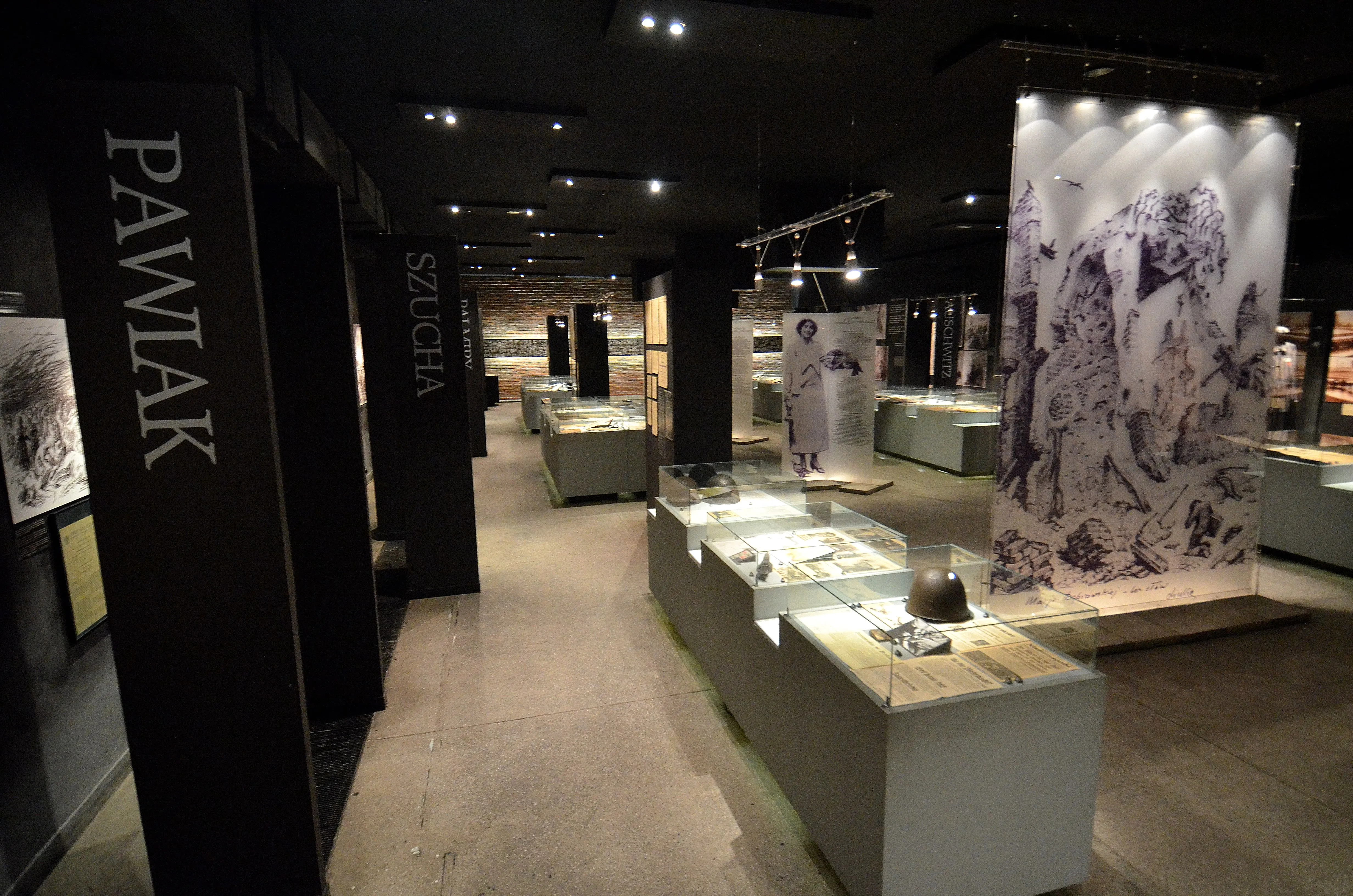Interior of Pawiak Prison Museum with display cases, a large artwork on a wall, and vertical signs with the name 'PAWIAK'