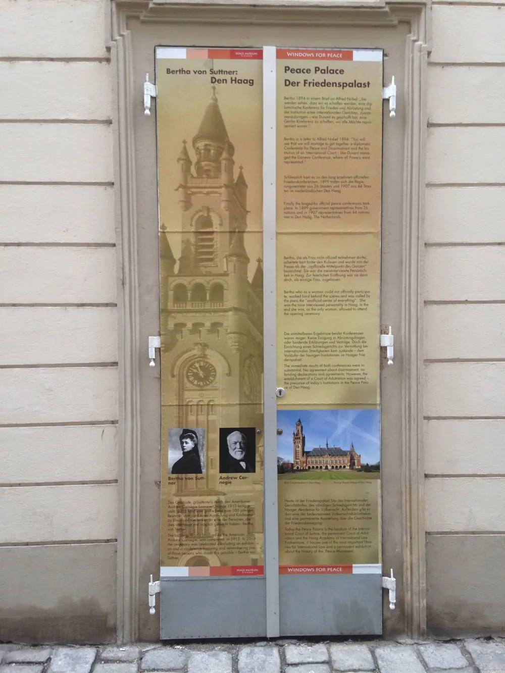 Buitendisplaypaneel voor het Vredesmuseum Wenen aan een gebouwmur, met tekst en afbeeldingen over het Vredespaleis en historische figuren.