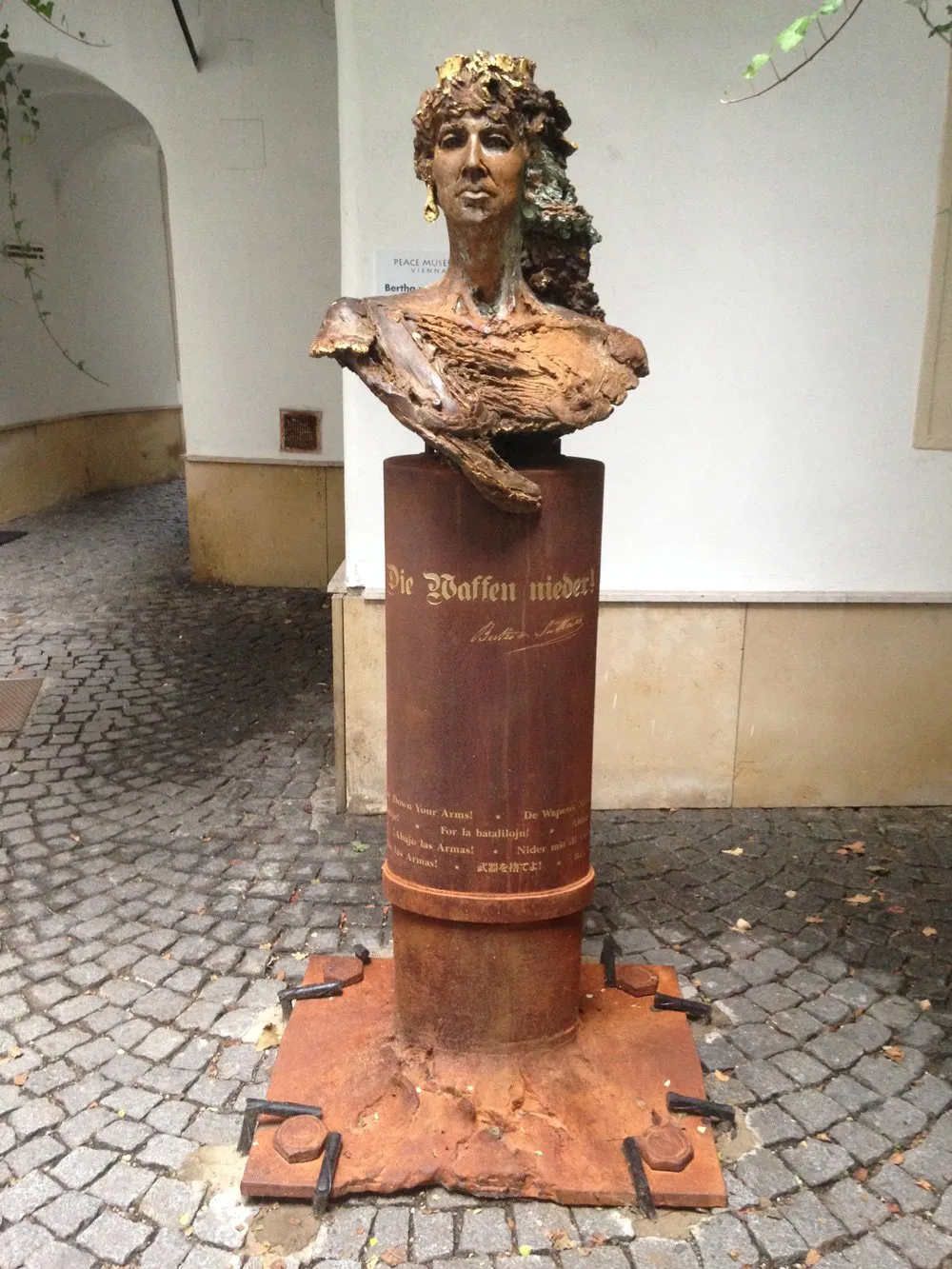 Bronzen bustesculptuur van een vrouw op een verroeste zuil met Duitse tekst, op een geplaveide binnenplaats