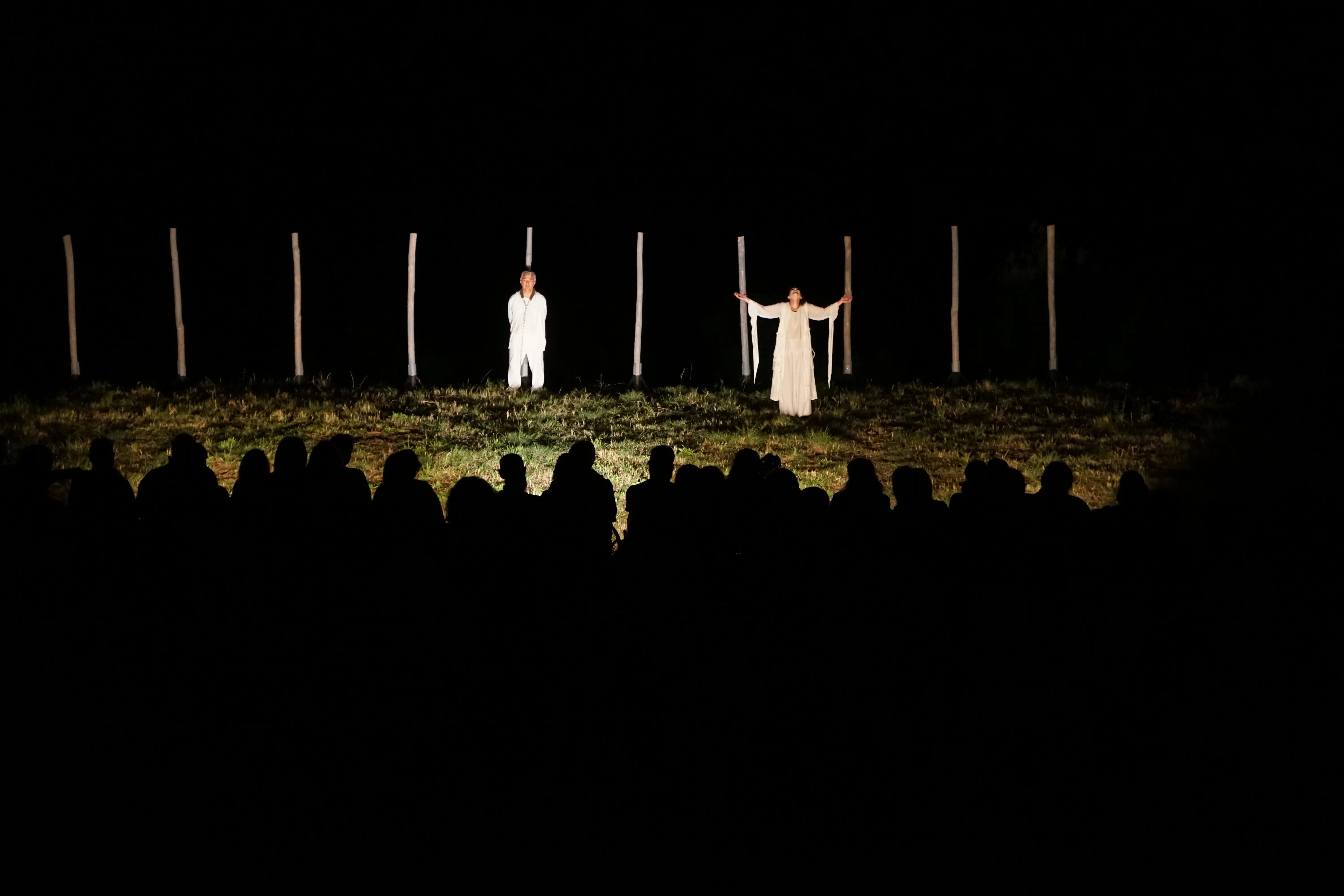 Dos artistas con trajes blancos en un escenario con poca luz y postes verticales al fondo, observados por una audiencia silueteada por la noche