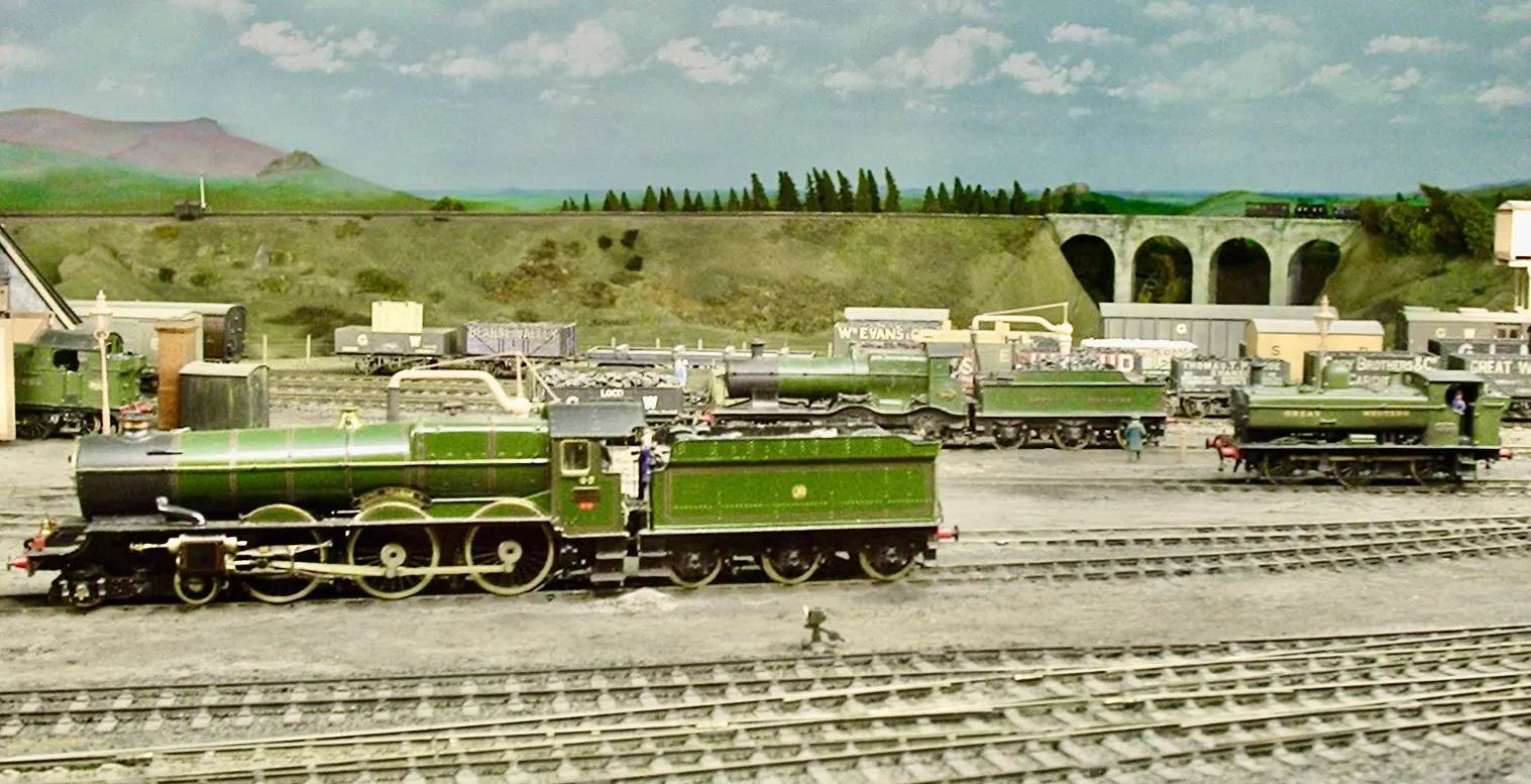 Exposición de trenes en miniatura con locomotoras de vapor verdes sobre múltiples vías en una detallada estación ferroviaria a escala con edificios y un viaducto de piedra frente a un telón de fondo de colinas