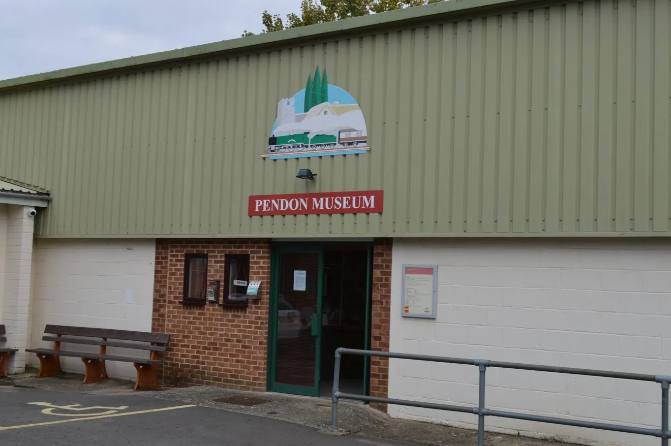 Vista exterior del Pendon Museum que muestra la fachada de metal corrugado verde con el letrero rojo PENDON MUSEUM, una puerta de entrada verde, bancos de madera y una rampa de acceso con barandillas metálicas.