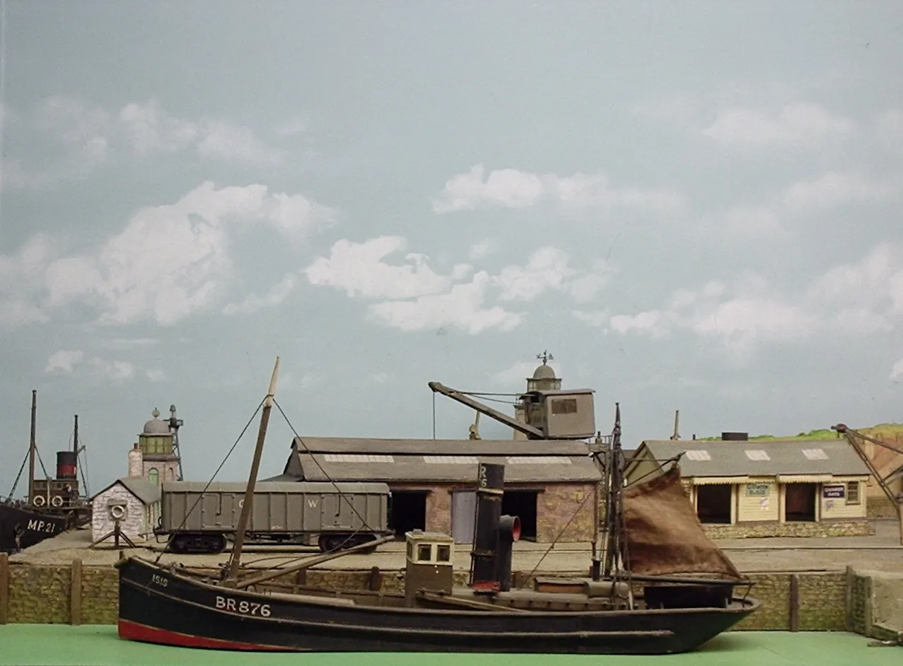 Detallado modelo en miniatura de una escena portuaria que presenta un barco pesquero negro con la etiqueta NR876, un faro, una grúa y edificios de piedra sobre un fondo de cielo pintado.