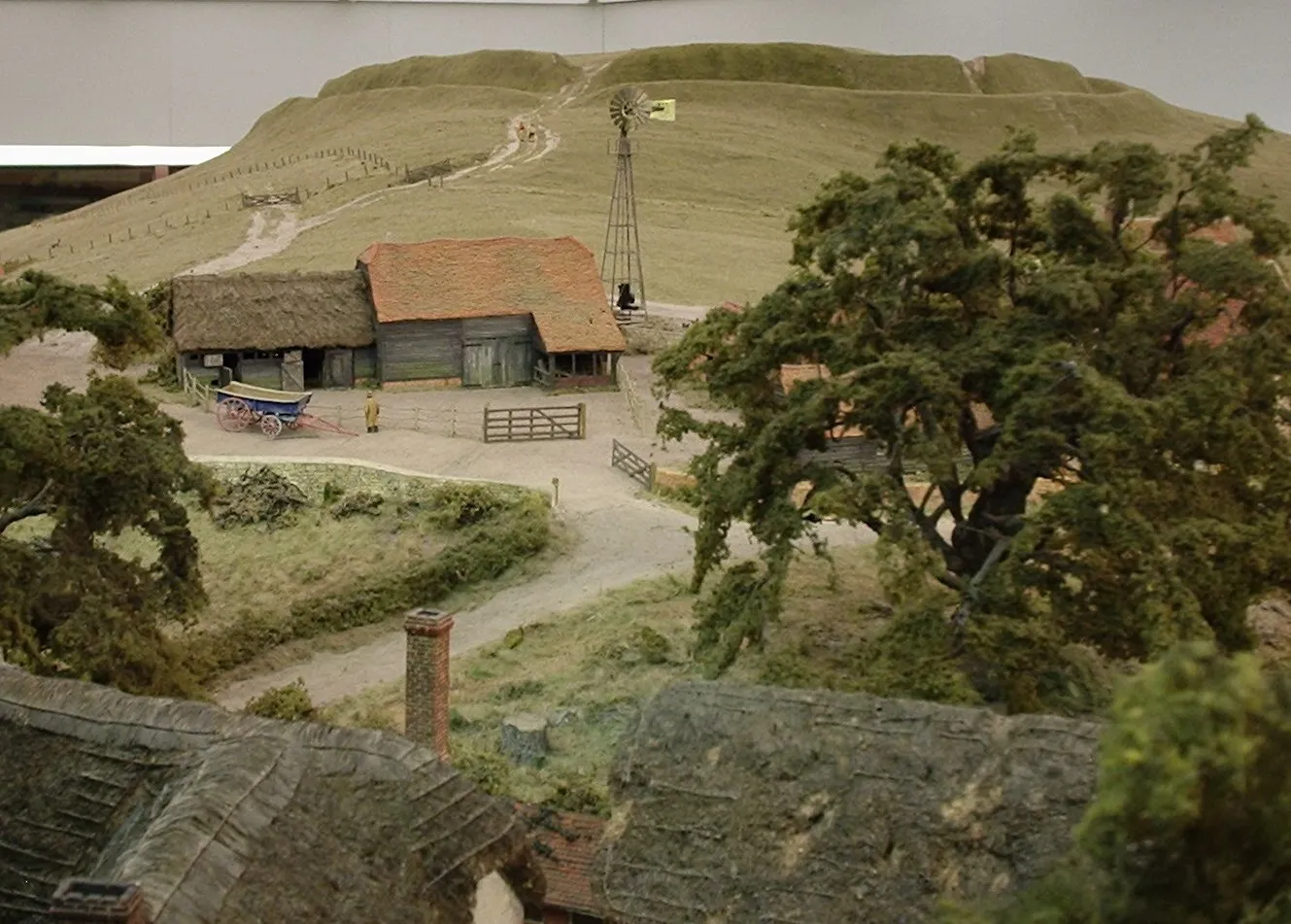 Maqueta de paisaje en miniatura que incluye una cabaña con techo de paja, un molino de viento, árboles y una colina con un sendero sinuoso