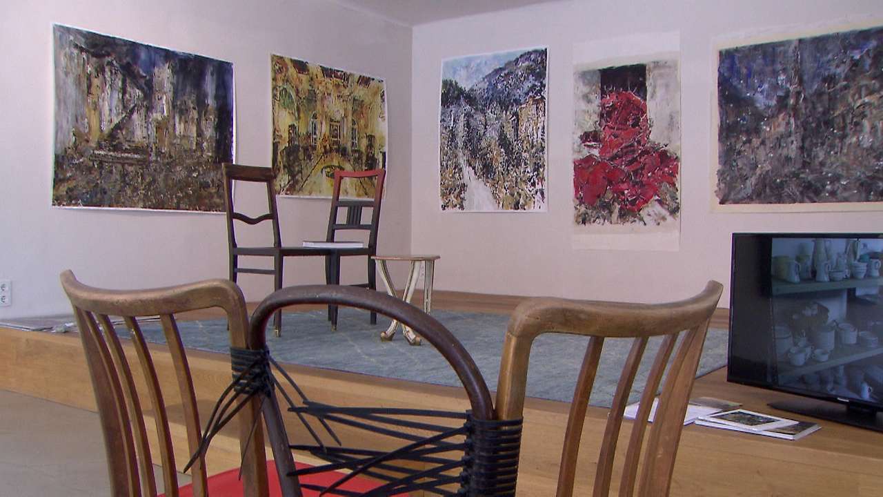 Interior de una galería con grandes pinturas abstractas en paredes blancas, sillas de madera dispuestas en el centro de la sala y un monitor de televisión en el lado derecho