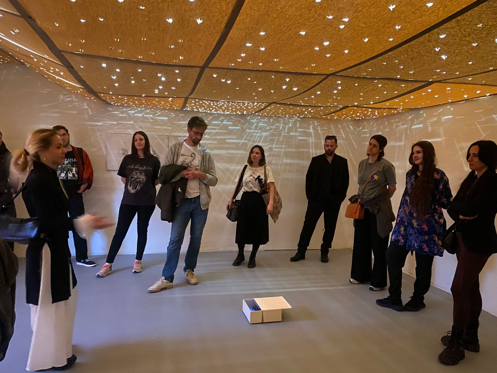 Los visitantes forman un círculo dentro de una galería con un techo cubierto de pequeñas luces, observando una pequeña caja en el suelo.
