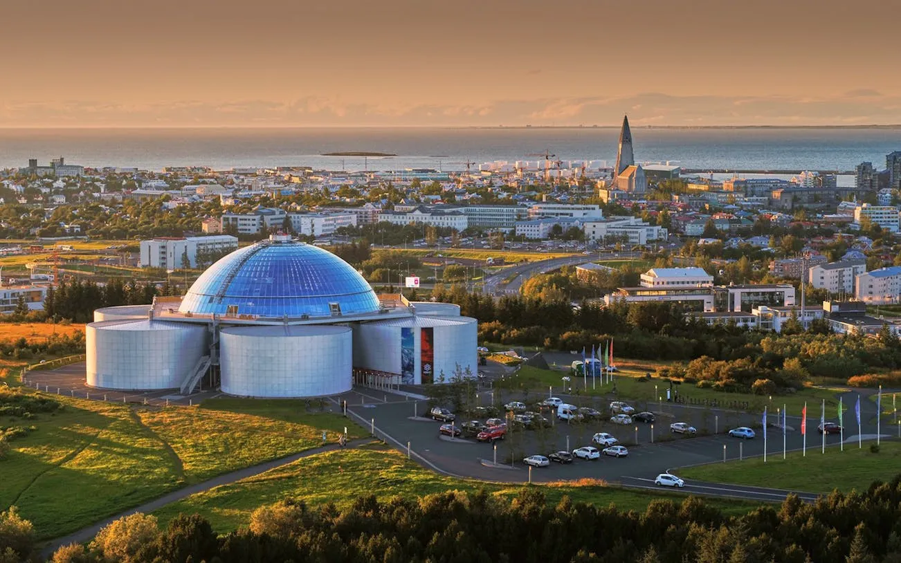 Luchtfoto van de kenmerkende blauwe glazen koepel en zilveren cilindrische tanks van Perlan, met het stadsgezicht van Reykjavík en de Hallgrímskirkja op de achtergrond bij zonsondergang.