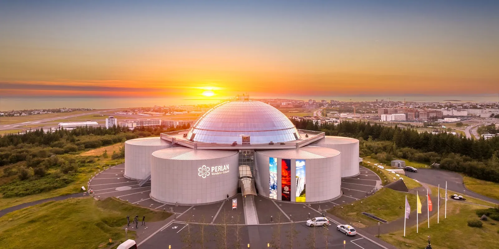 Panoramisch luchtfotografisch beeld van het Perlan-museum, met een grote glazen koepel boven op witte cilindrische tanks tijdens een levendige zonsondergang boven Reykjavík.