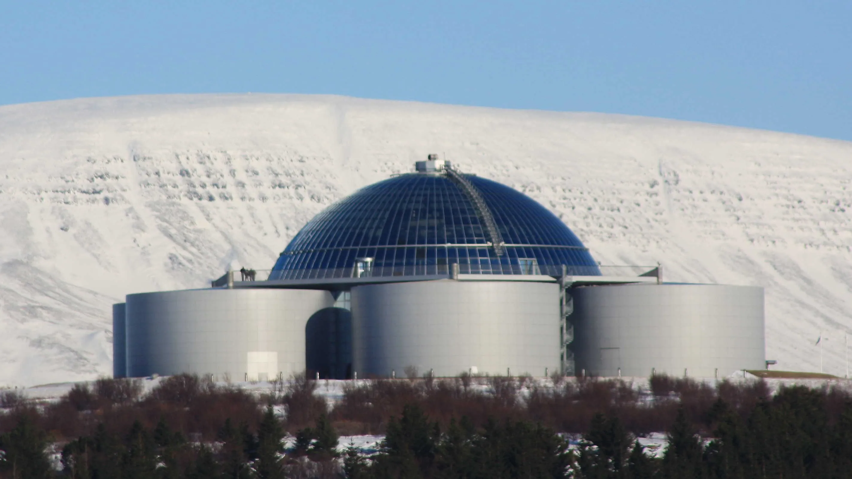 Het Perlan-gebouw met zijn grote glazen koepel boven op cilindervormige zilveren tanks, gelegen tegen een met sneeuw bedekte heuvel onder een helderblauwe lucht.