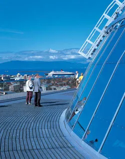 Twee mensen staan op het gebogen houten uitkijkplatform van Perlan, met de blauwe glazen koepel aan de rechterkant en een panoramisch uitzicht op de haven van Reykjavík en de bergen op de achtergrond.