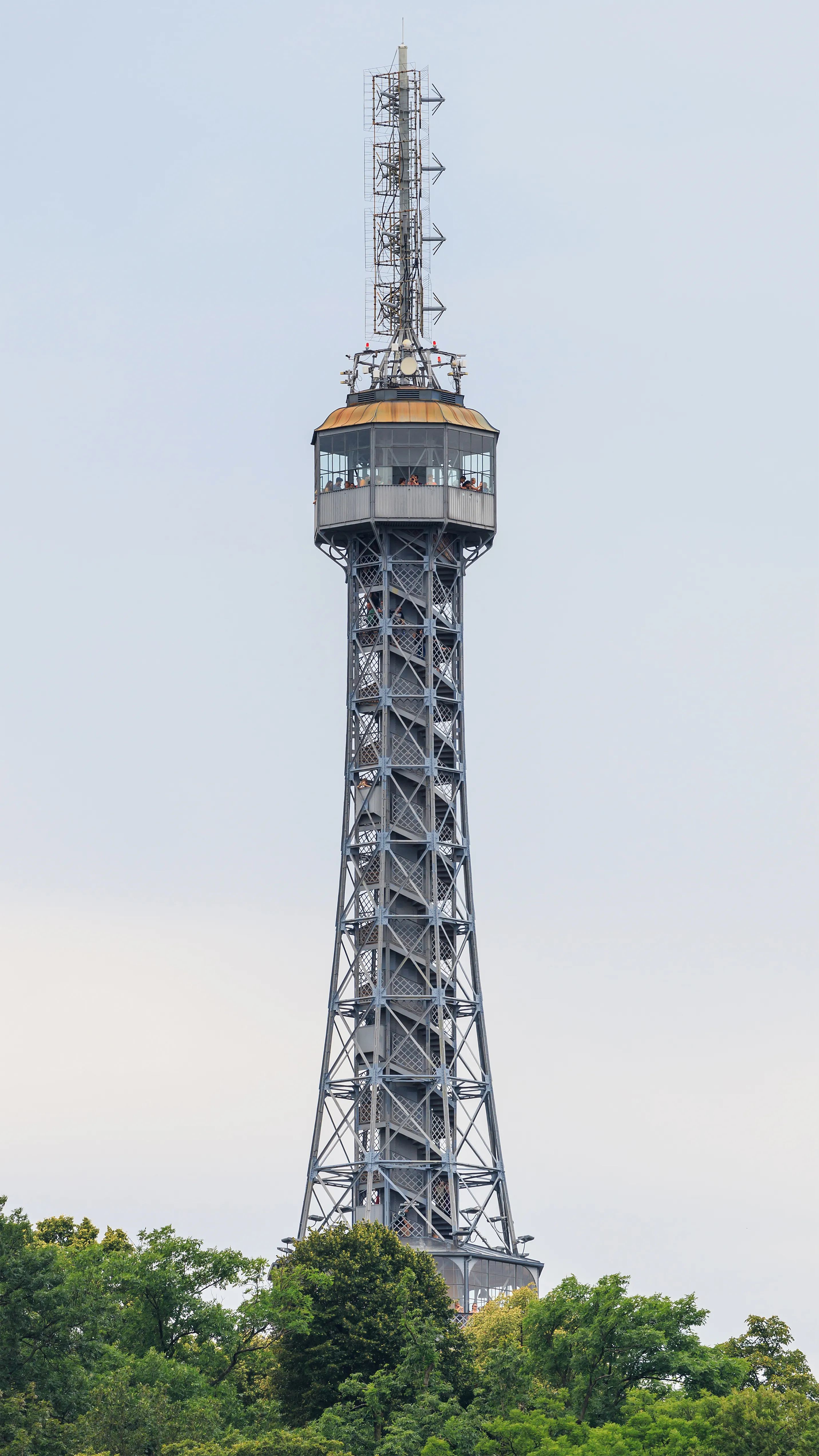 Hoge stalen observatietoren met een vakwerkstructuur en observatiedek, omgeven door groene bomen onder een lichte lucht