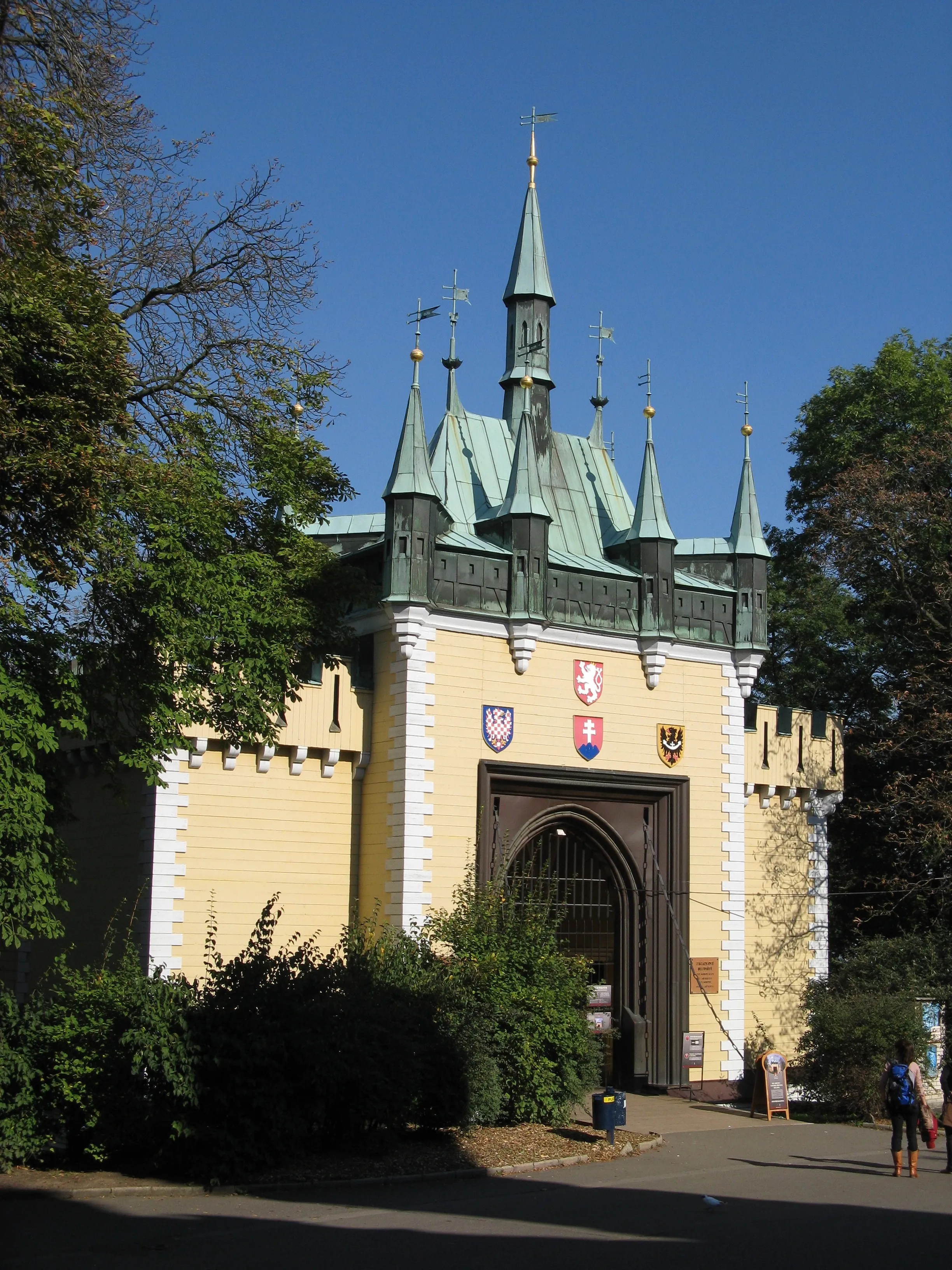 Gele en witte neogotische paviljoen met groene torenspitsen en heraldische schilden, toegangspoort, omringd door bomen en struiken onder een helderblauwe hemel