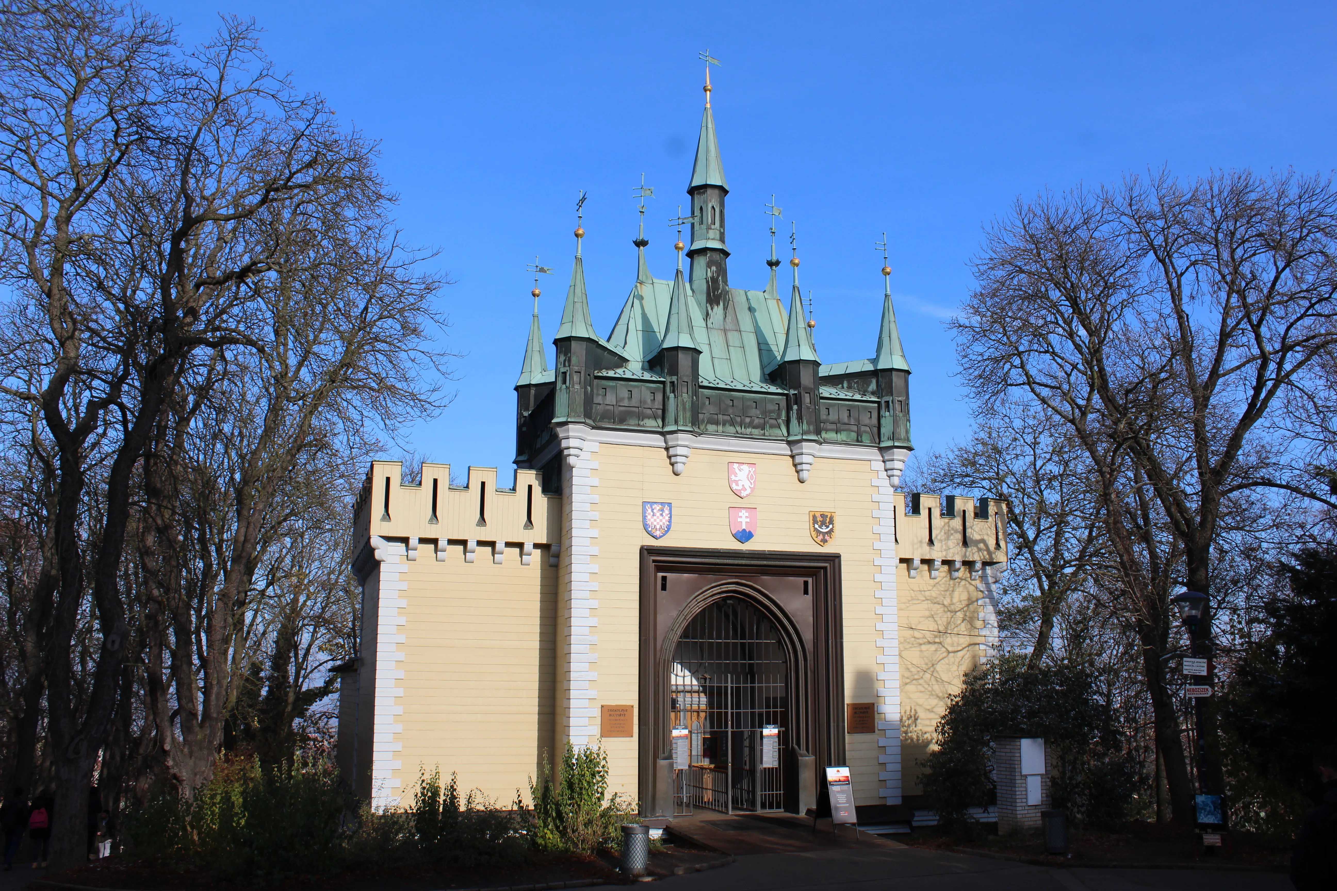 Een geel gebouw met groene torenspitsen en een ingang met een spitsboog, omringd door kale bomen onder een blauwe hemel