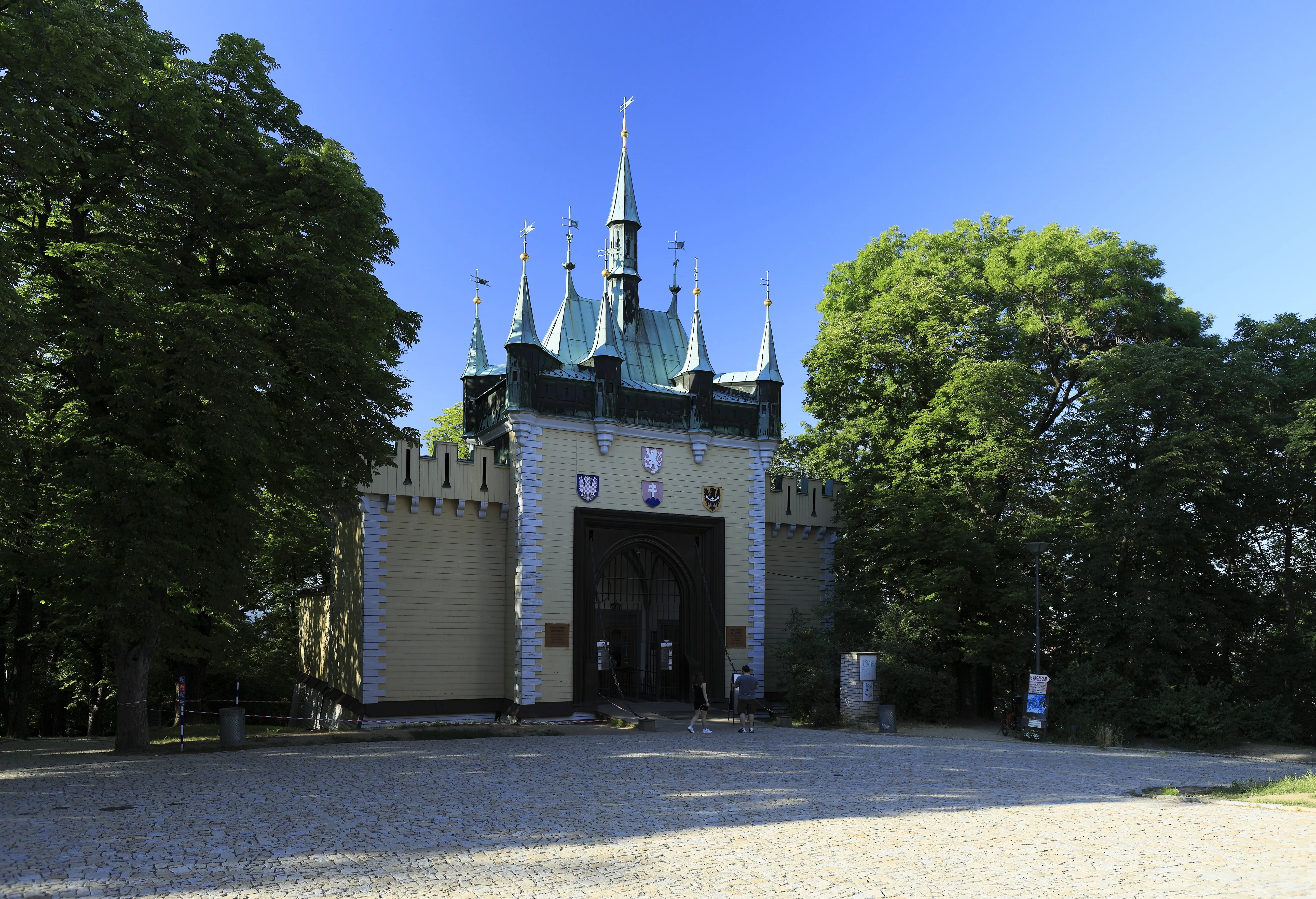 Een mock-Gotisch paviljoen met groene torenspitsen en een centrale boogpoort, omringd door bomen onder een helderblauwe lucht.