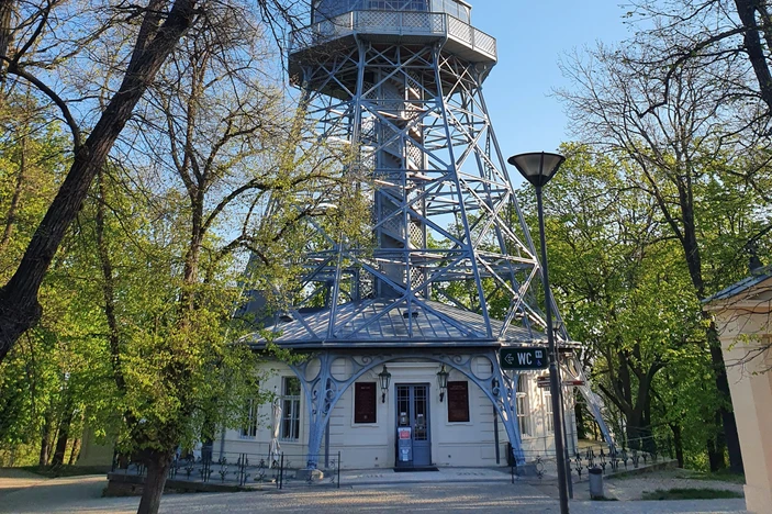 Een wit gebouw met een blauwe metalen torenstructuur op de Petřín-heuvel, omringd door groene bomen en een lantaarnpaal op de voorgrond.