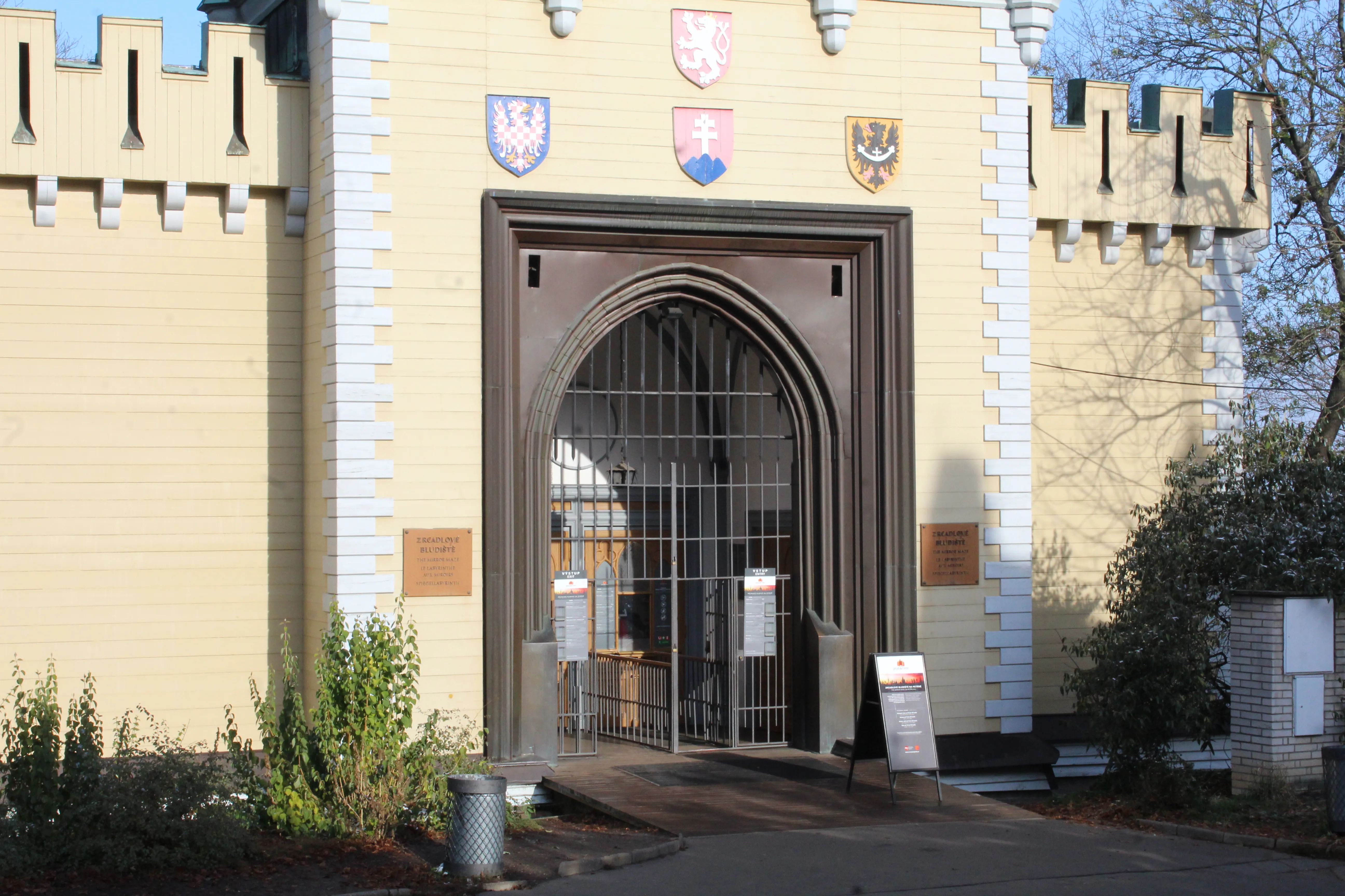 Geel neogotisch gebouw met gekanteelde muren, een boogvormige ingang met een metalen hek, en drie heraldische schilden boven de deur