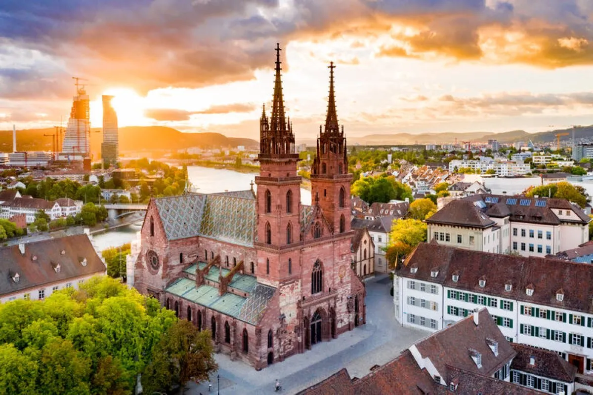 Aerial view of Basel Minster's twin red sandstone spires at sunset with the Rhine river and city skyline glowing in warm golden light