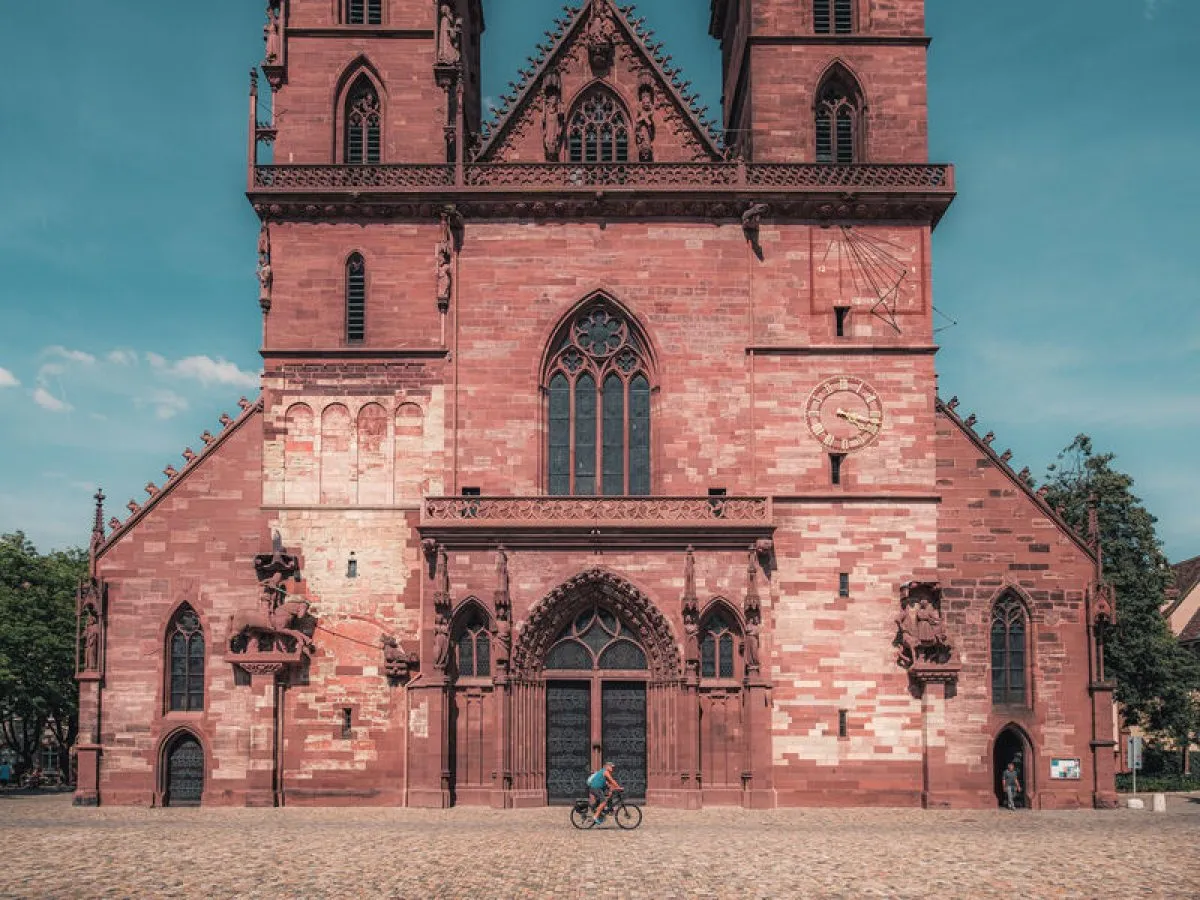 Rear facade of Basel Minster, a red sandstone Gothic cathedral with twin towers and arched windows, with a cyclist on the cobblestone square in front.