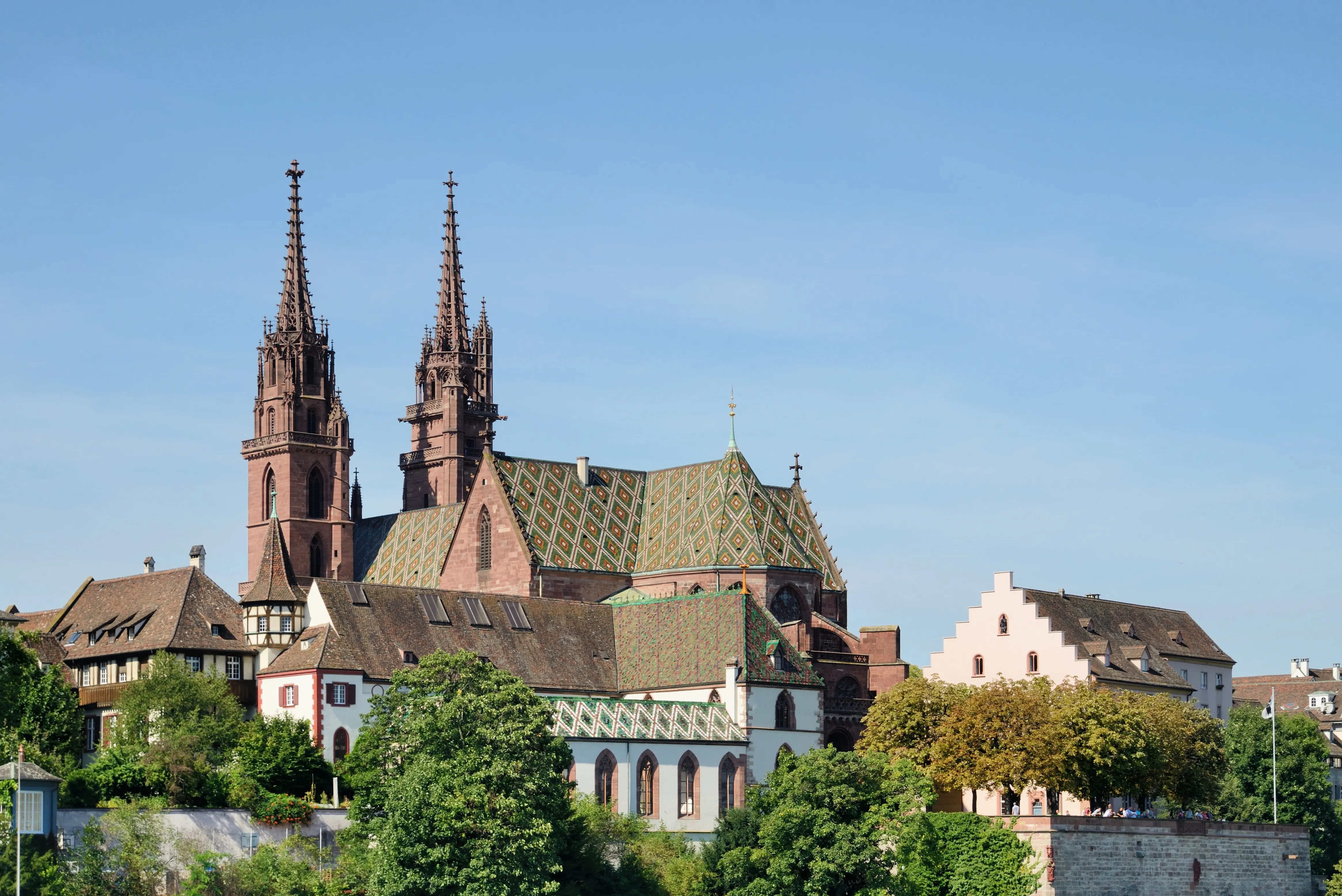 Basel Minster cathedral featuring twin gothic red sandstone spires and a green patterned tile roof, surrounded by historic buildings and green trees under a clear blue sky.