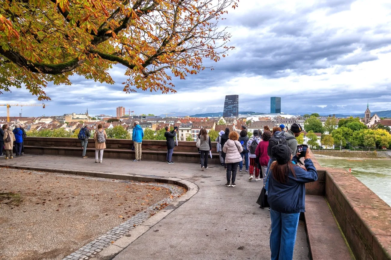 People standing on a curved stone terrace overlooking Basel's cityscape with autumn tree branches overhead and the Rhine river visible to the right
