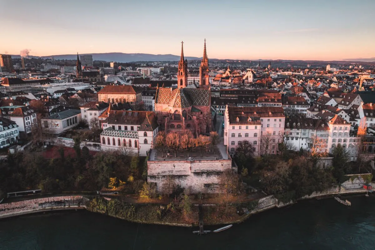 Aerial view of Basel Minster cathedral with its twin spires overlooking the Rhine river at sunset, surrounded by the old town rooftops