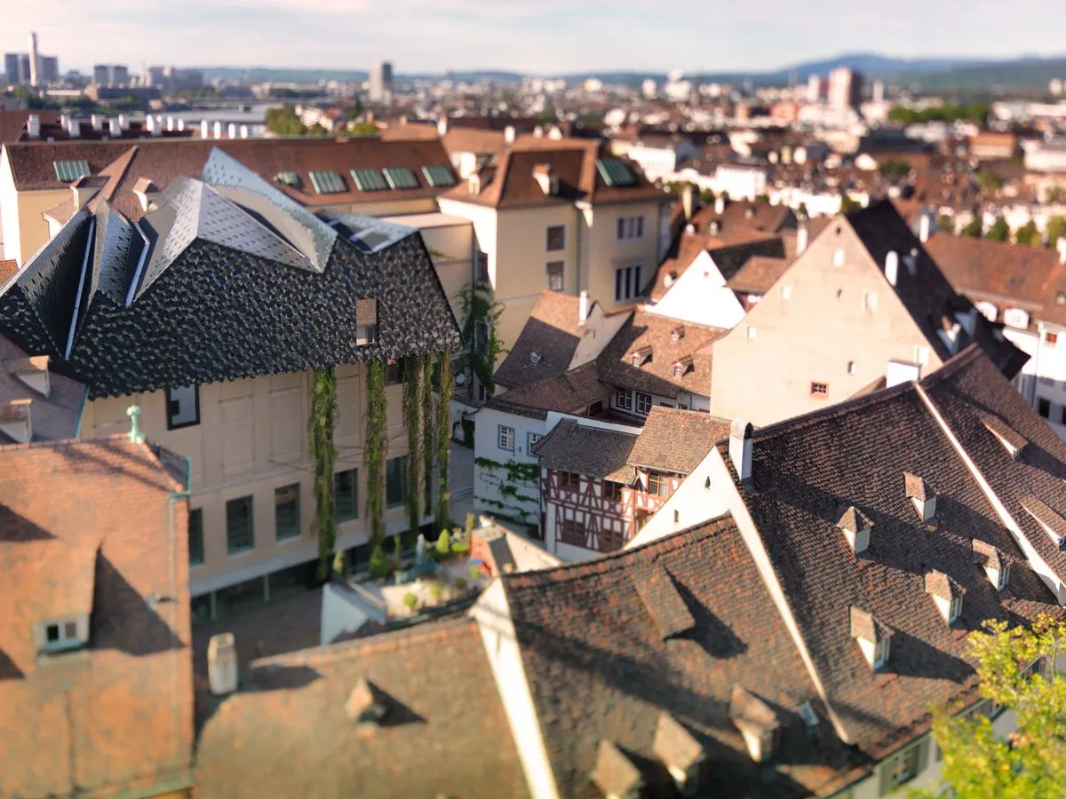 Aerial tilt-shift view of Basel's old town rooftops showing traditional European architecture with terracotta tiles and a modern building with a dark angular roof and climbing plants, with the cityscape stretching into the distance.