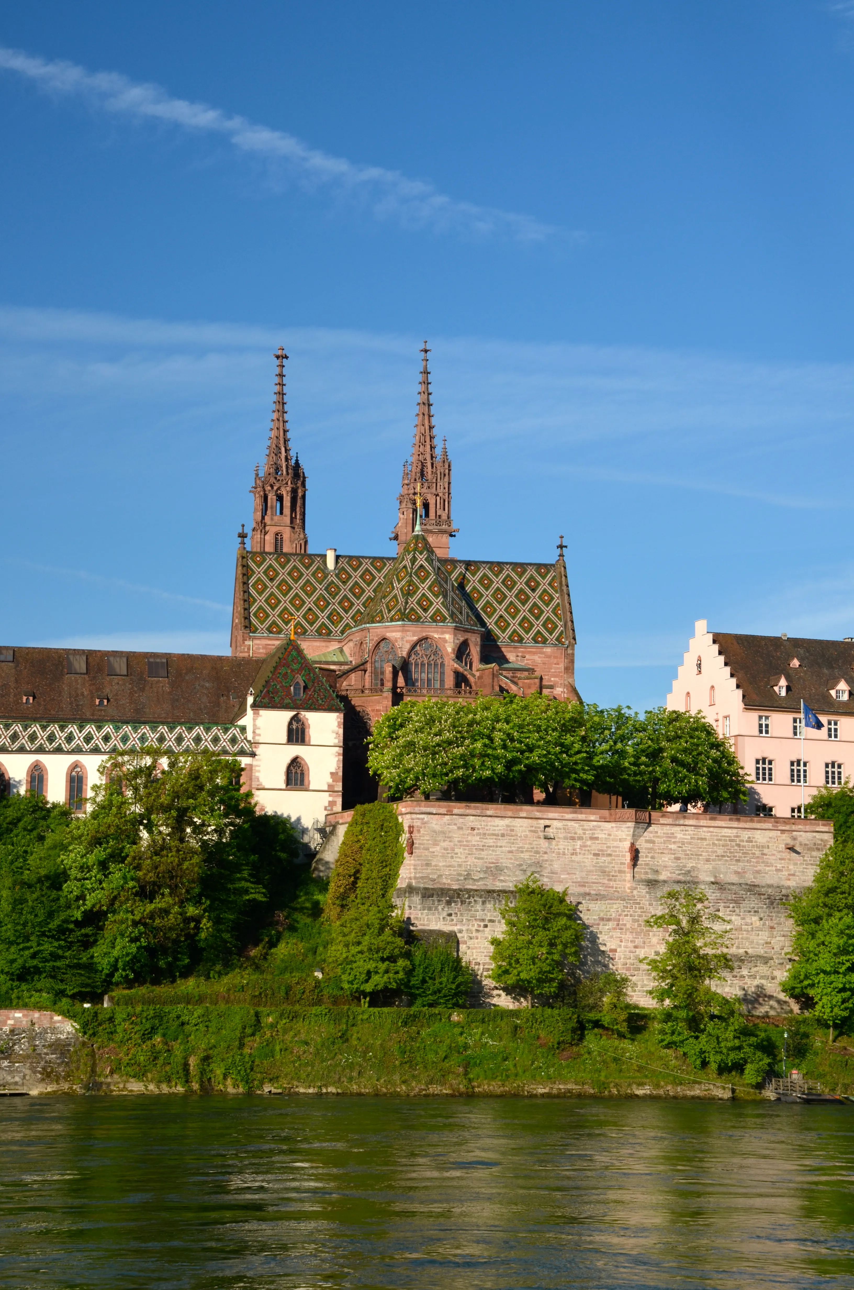 Basel Minster cathedral with twin spires and colorful tiled roof overlooking the Rhine river, with stone terrace walls and green trees.