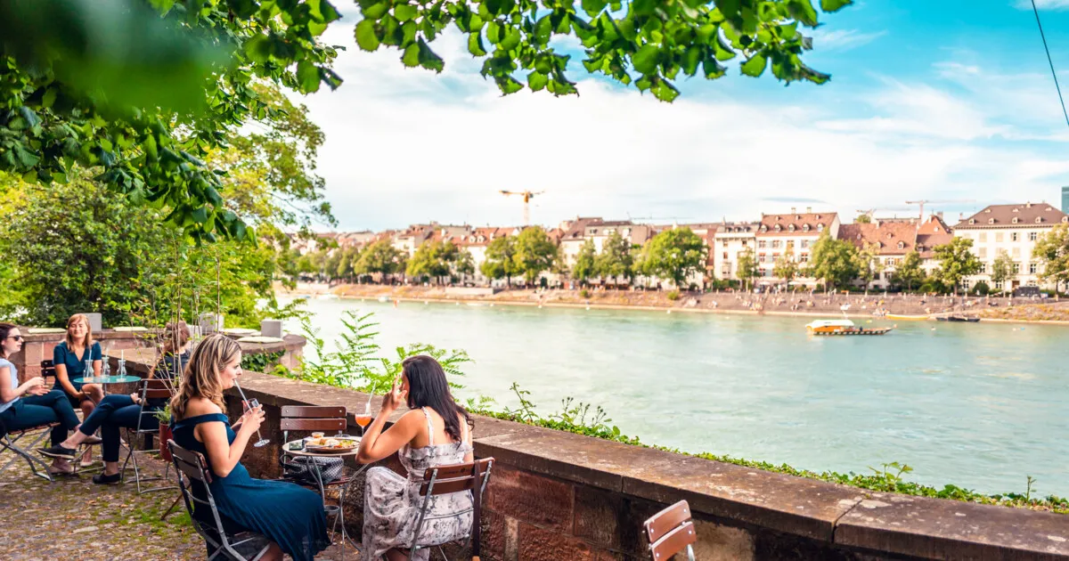People sitting at tables on a sunny stone terrace overlooking the Rhine river and the city of Basel, framed by green trees.
