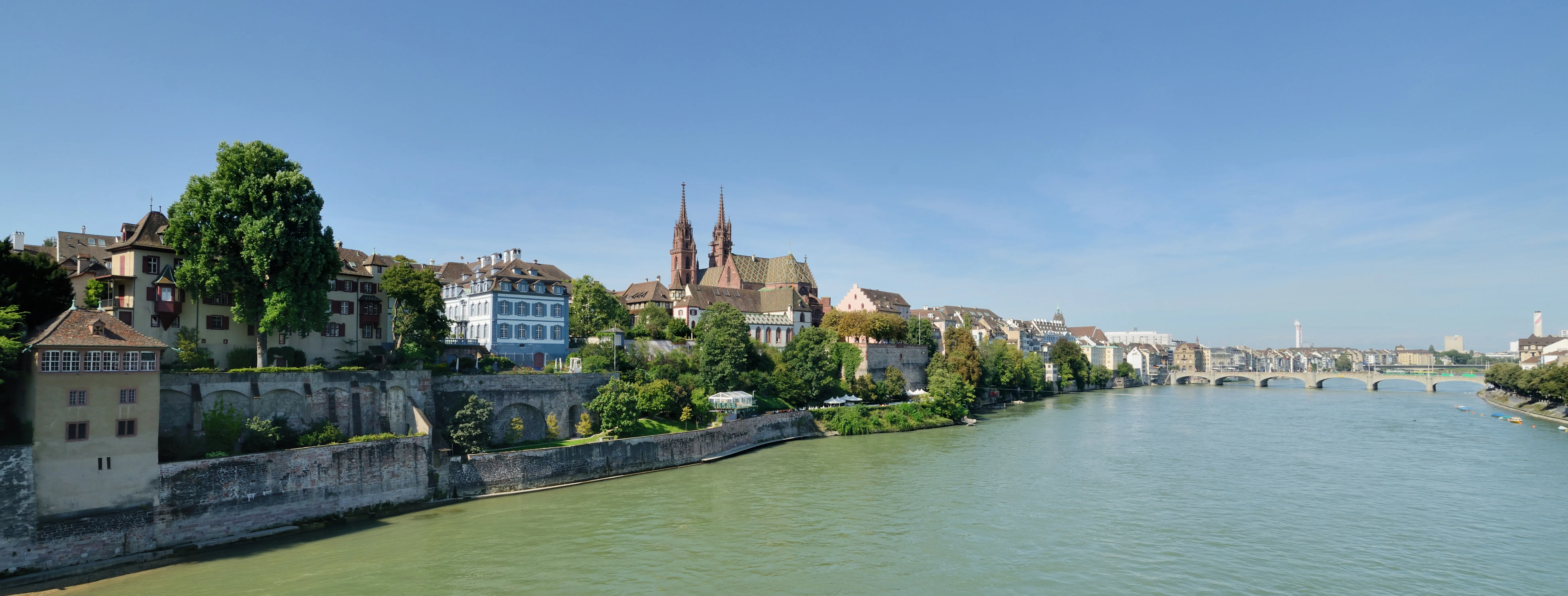 Panoramic view of the Rhine river in Basel with the twin towers of Basel Minster and historic buildings along the riverbank under a clear blue sky.