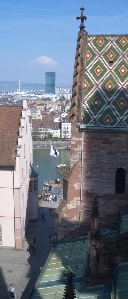Aerial view of Basel Minster's colorful diamond-patterned tiled roof and stone tower with cross, overlooking the Rhine river and Basel cityscape with the Roche Tower visible in the background